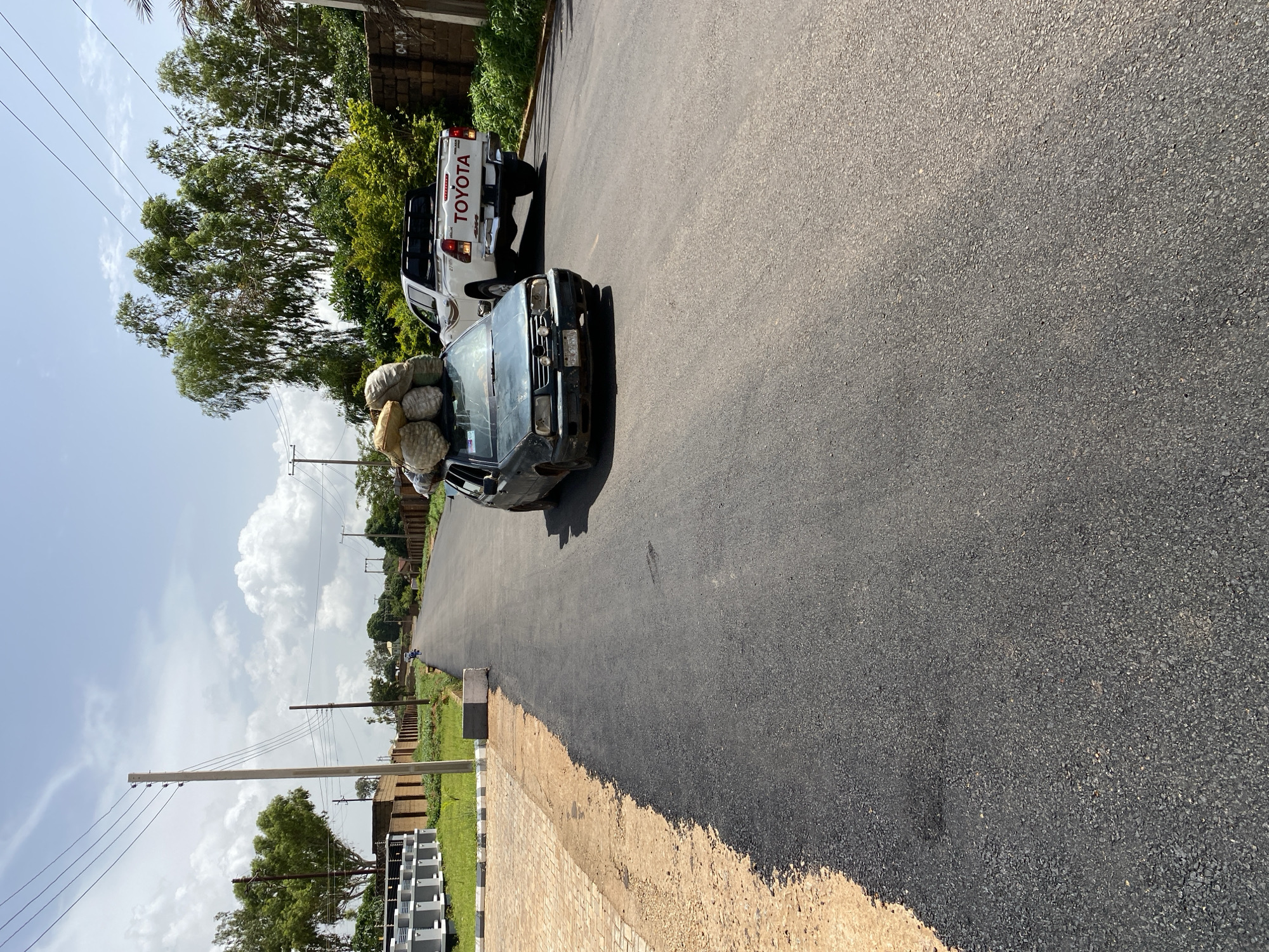 In Bukuru, Nigeria, on a freshly paved asphalt road, a dark green sedan carrying a significant load of sacks piled high on its roof travels forward. Following closely behind is a white Toyota pickup truck, also moving in the same direction. The road is flanked by a newly constructed tiled sidewalk on the left and a dirt shoulder. Beyond the sidewalk, modern buildings with gray structures are visible, along with a fence. On the right side of the road, there is lush green vegetation and trees. Power lines stretch across the sky, supported by utility poles. The sky is a mix of blue with scattered white clouds, indicating a daytime scene.