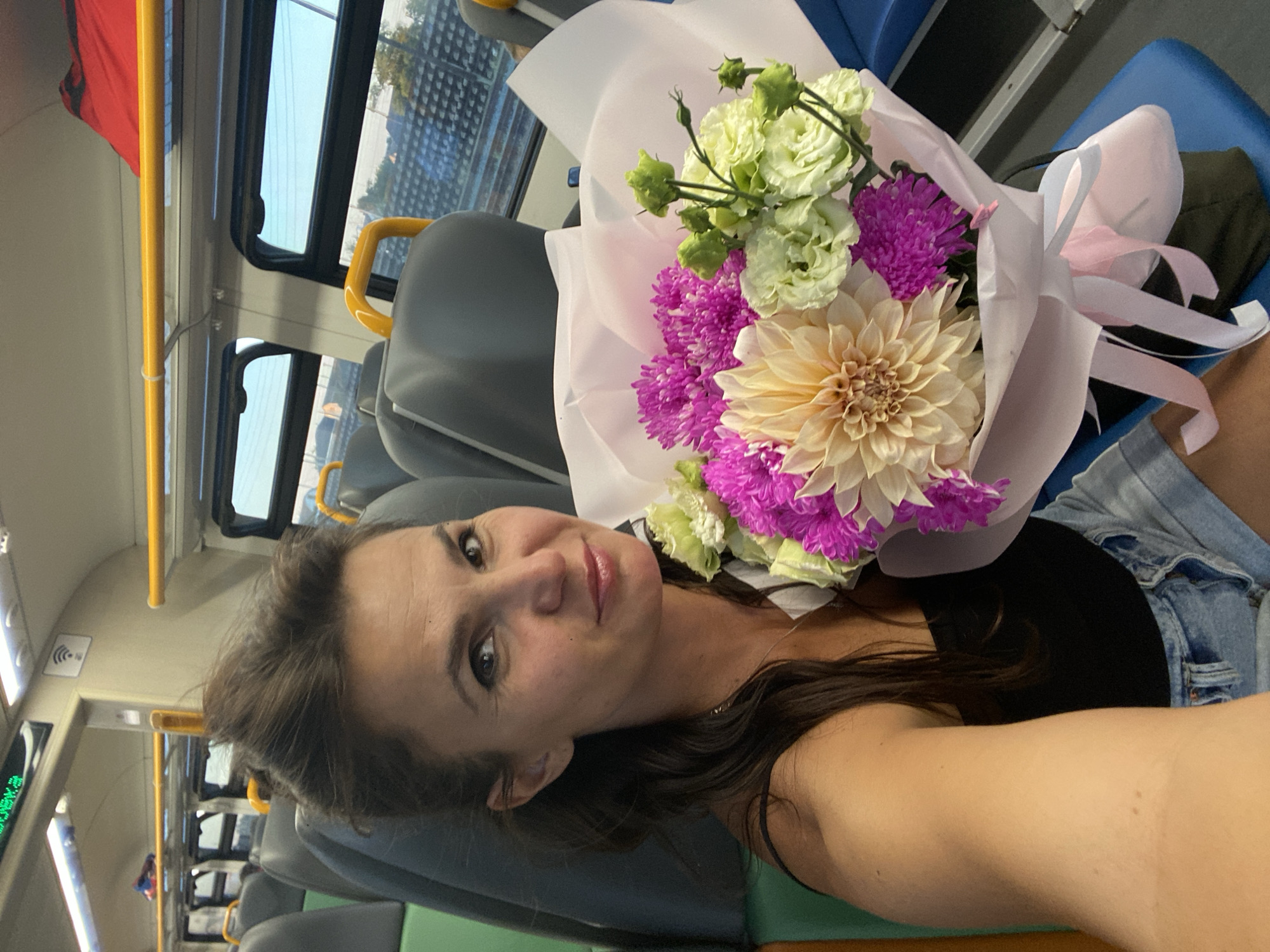 A woman is holding a bouquet of flowers while sitting on a train. She has brown hair and is wearing a black tank top and denim shorts. The train appears to be modern with grey seats and yellow handrails. The windows show a view of the landscape outside, which includes trees and buildings. The bouquet consists of a large beige dahlia, several pink chrysanthemums, and a few light green roses. The flowers are wrapped in pink and white paper and tied with a pink ribbon. The woman is looking at the camera and smiling. The image is a selfie taken by the woman.