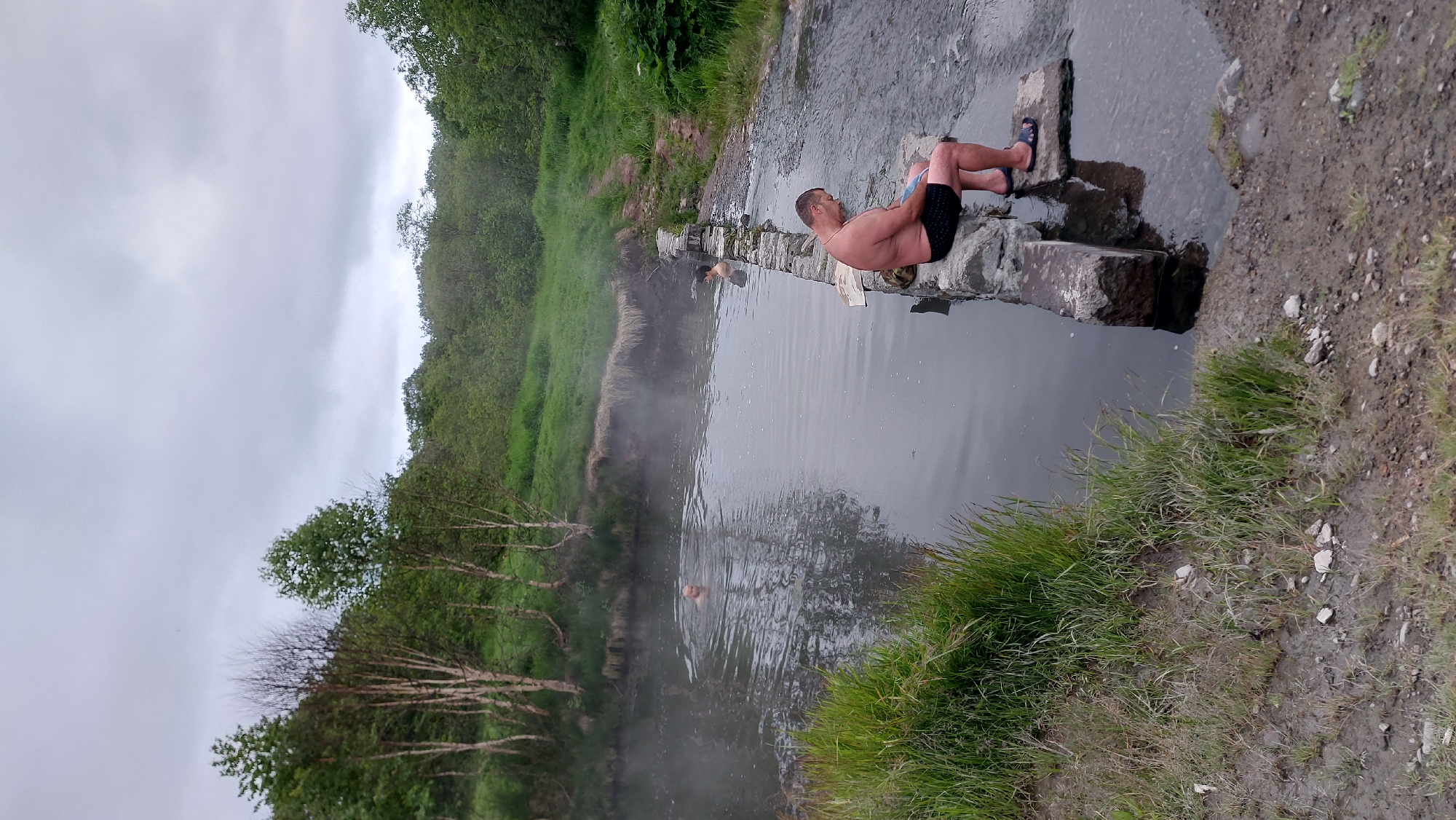 A man sits on a stone edge of a hot spring in Nachiki, Russia, with his legs submerged in the water. He is wearing black shorts and blue sandals. The hot spring is murky and steaming, surrounded by lush green vegetation. In the background, another person is partially visible in the water. The sky is overcast, creating a diffused light. The ground around the spring is muddy with scattered rocks.