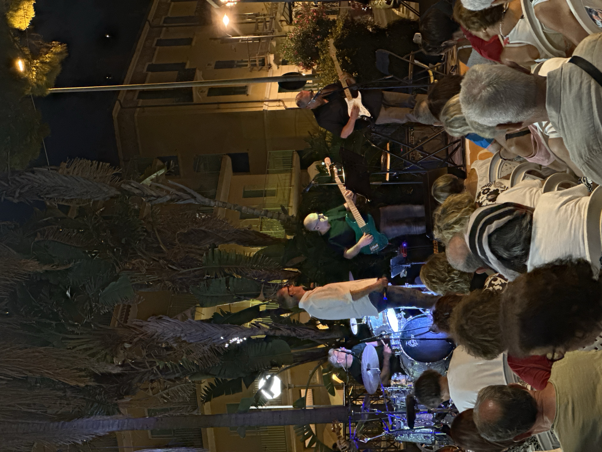 This is an outdoor evening scene in Ospedaletti, Italy, likely on a street or a small piazza, with a band performing. A group of musicians, consisting of a drummer, two guitarists, and possibly a singer, are on a raised platform. The drummer is seated behind a drum kit, one guitarist is playing a green electric guitar, and another is playing a white electric guitar. A singer, wearing a light-colored shirt, stands center stage. In the foreground, an audience is seated in chairs, facing the stage, with their backs to the camera. The background shows a building with balconies and windows, illuminated by streetlights and lights from the performance. Palm trees and other foliage are visible, adding to the Mediterranean atmosphere. The overall impression is of a casual, local musical event.