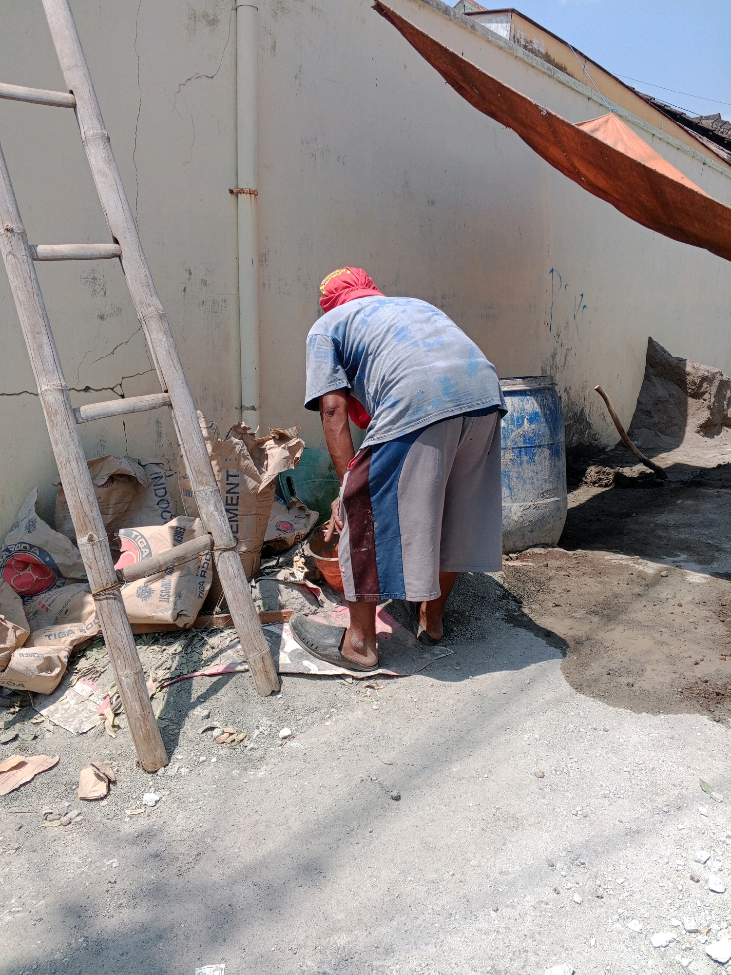 In Giritirto, Indonesia, a construction worker, wearing a blue t-shirt and gray shorts with a red bandana, is seen from the back tending to a pile of cement bags. A bamboo ladder is positioned to the left of the cement bags, which appear to be made of brown paper with some text. A large blue barrel stands to the right of the worker, and a wooden hoe lies on the ground in the background. The environment is dusty, with a concrete wall and a reddish-orange tarp in the upper right corner, suggesting an outdoor construction or renovation site under a clear sky.