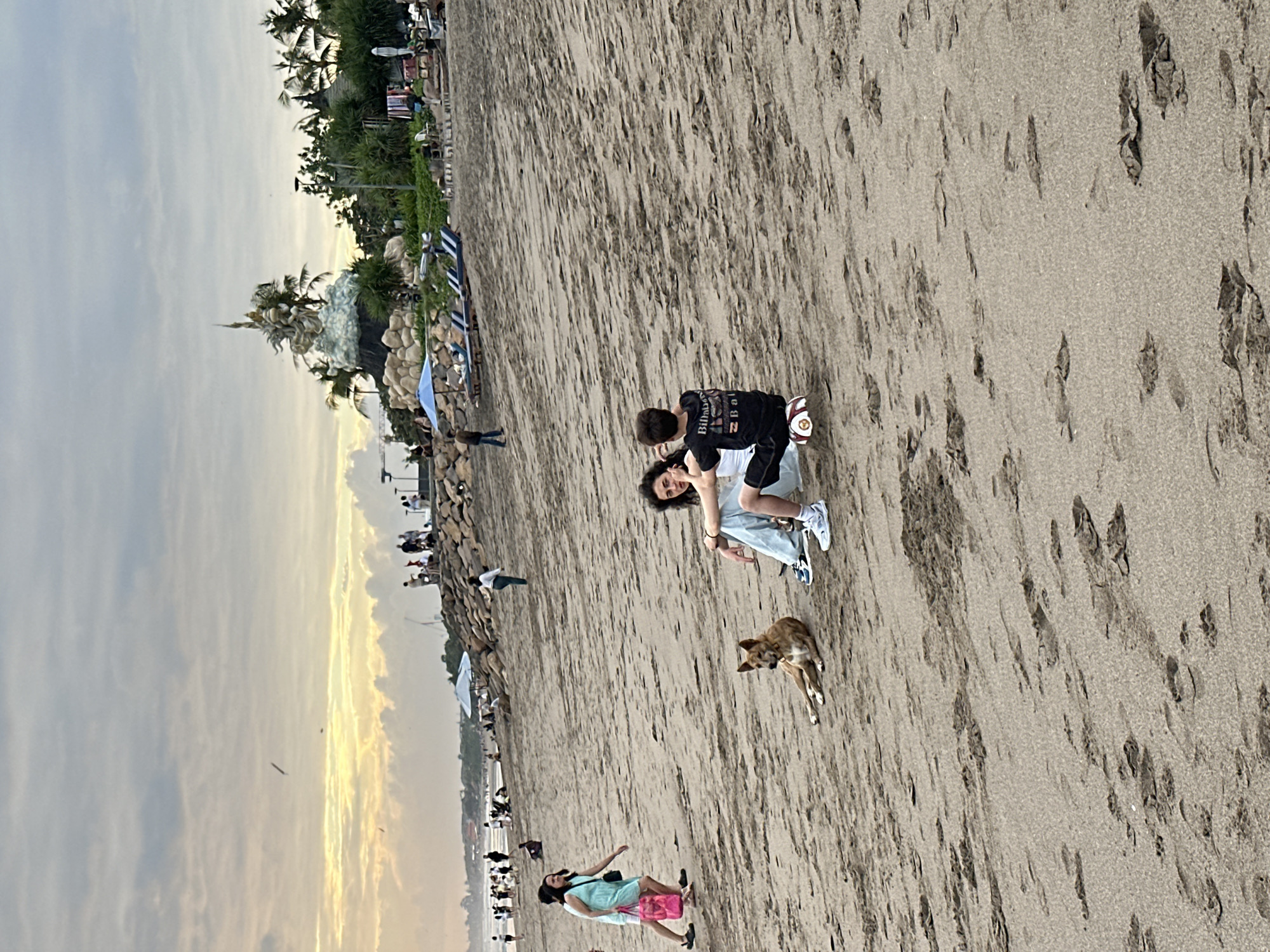 In North Kuta, Indonesia, a beach scene unfolds under a cloudy sky with hints of sunset. In the foreground, two individuals, one seated and facing away, the other sitting beside them, interact. A brown dog lies relaxed on the sand between them. To the left, a woman walks with a pink bag. In the mid-ground, several people are visible scattered across the beach, with some walking near the water's edge. Beach chairs and umbrellas are set up further back, near a rocky embankment adorned with palm trees and a large, ornate sculpture. The sand is textured with numerous footprints.
