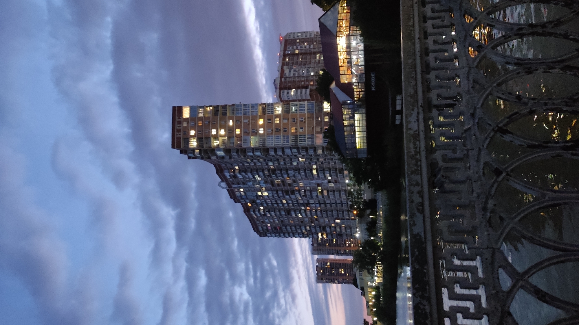 This is a nighttime exterior shot of a waterfront area in Krasnodar, Russia. In the background, several tall, modern apartment buildings are illuminated by interior lights. The building in the foreground has a distinctive curved facade and is significantly taller than the others. Adjacent to this building is a low-rise structure with large glass windows, also lit from within, displaying the Cyrillic text "КАФЕ" (cafe) above its entrance. The scene is framed by a decorative metal railing in the foreground, which is positioned over a body of dark water reflecting the lights from the buildings and the sky. The sky above is a deep twilight blue, filled with dramatic, dark clouds that are beginning to clear, hinting at the end of the day. The overall impression is one of an urban evening landscape with residential and commercial elements near a body of water.