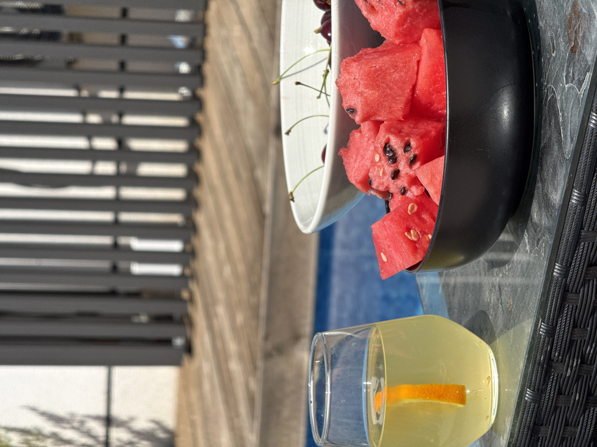 In Lastva Grbaljska, Montenegro, a glass of lemonade with an orange slice sits on a glass-topped table. Next to it, a black bowl overflows with cubed watermelon, and a white bowl behind it contains a scattering of cherries. The background features a blurred swimming pool and a wooden deck, suggesting an outdoor recreational setting. A grey fence is visible further in the background. The lighting indicates a sunny day.