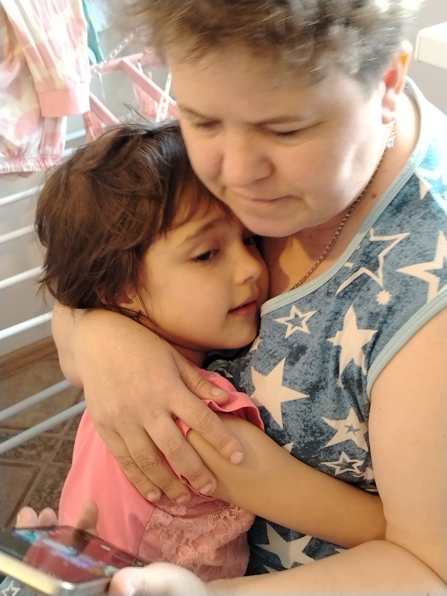 This image, taken in Noviy, Russia, captures an intimate moment between an adult and a child in what appears to be a domestic indoor setting. The adult, with short, light brown hair, is positioned on the right, gently embracing the child. She is wearing a blue sleeveless top adorned with numerous white stars and a silver chain necklace. Her gaze is directed downwards, suggesting a tender focus on the child or something they are jointly observing.
The child, with dark brown hair, is nestled into the adult's embrace. They are wearing a pink top, possibly with lace detailing, and are looking towards the left of the frame. The child's hands are visible at the bottom of the image, holding a smartphone, implying they are engaged with the device, possibly showing something to the adult or watching content.
The background is softly blurred but reveals elements of a home environment. To the upper left, a pink or white clothes drying rack is visible, with a pink and white patterned garment hanging on it. The floor beneath the subjects appears to be tiled with a geometric pattern. The lighting suggests it is daytime. The image conveys a sense of comfort, affection, and shared attention between the two individuals. There is no visible text in the image.