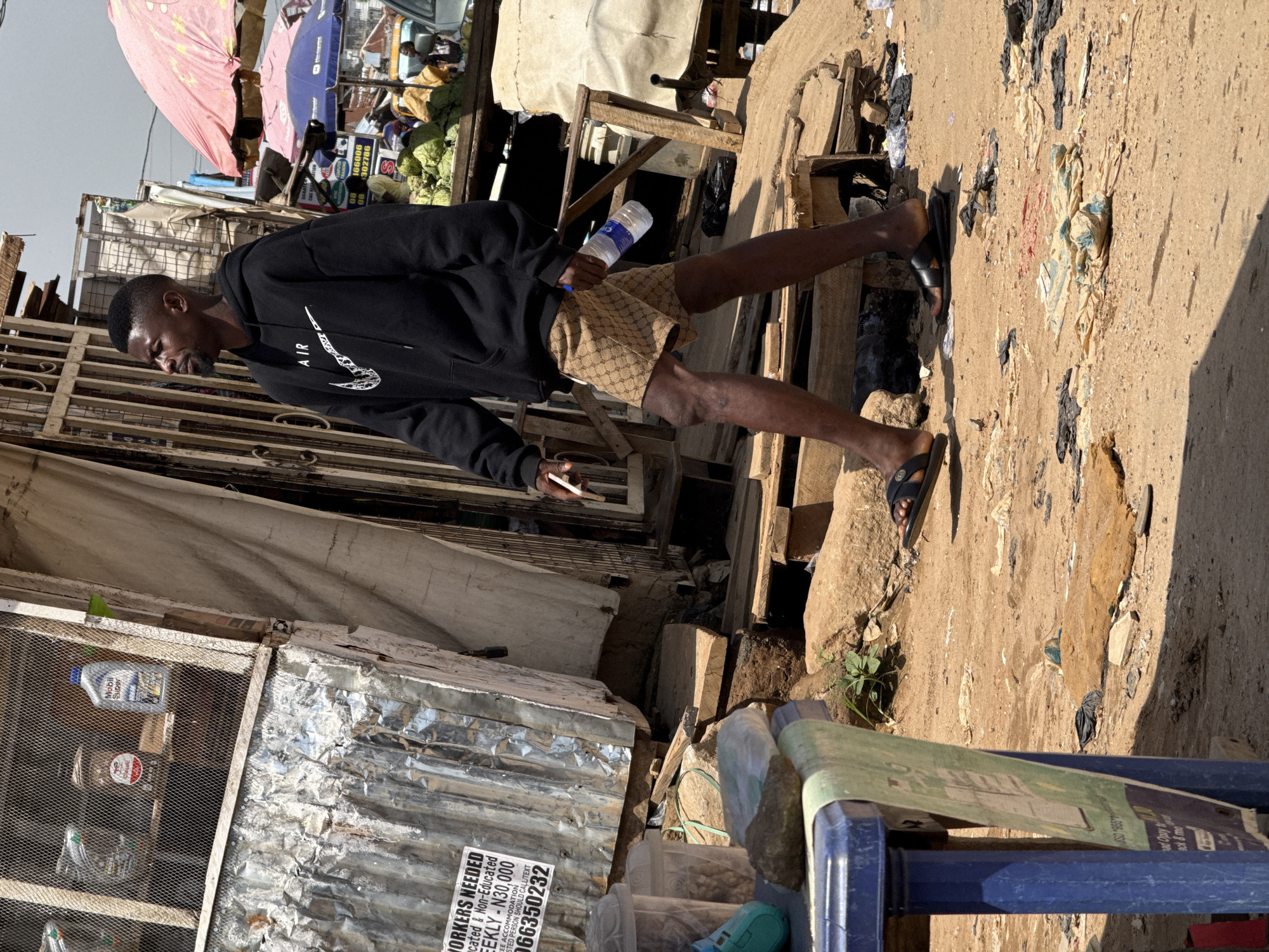 A man in a black Nike hoodie and patterned shorts walks down a dusty street in Abuja, Nigeria, holding a bottle of water. He is walking past a market stall with a corrugated metal exterior, displaying products behind a wire mesh. The ground is covered in sand, debris, and discarded plastic. In the background, other market stalls and people are visible, along with umbrellas providing shade. The overall scene depicts a busy, outdoor market environment.