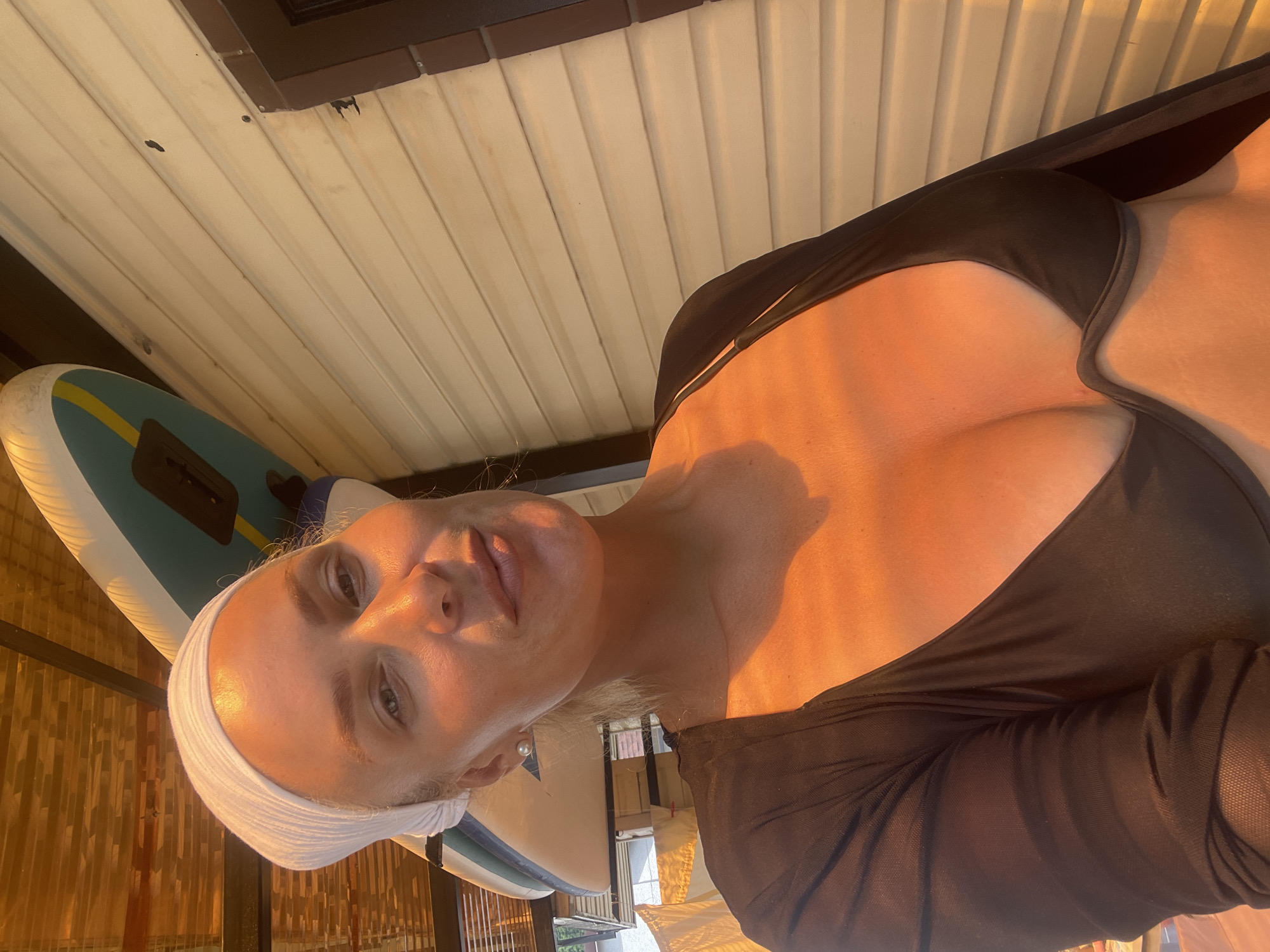 A blonde woman wearing a white headscarf and a black swimsuit top is pictured outdoors. She is positioned close to the camera, with a light-colored siding wall behind her. Above and to her left, two paddleboards are visible, leaning against a structure with a slatted roof. The setting appears to be a sunny outdoor area, possibly near water given the presence of the paddleboards. The woman is looking directly at the camera with a slight smile.