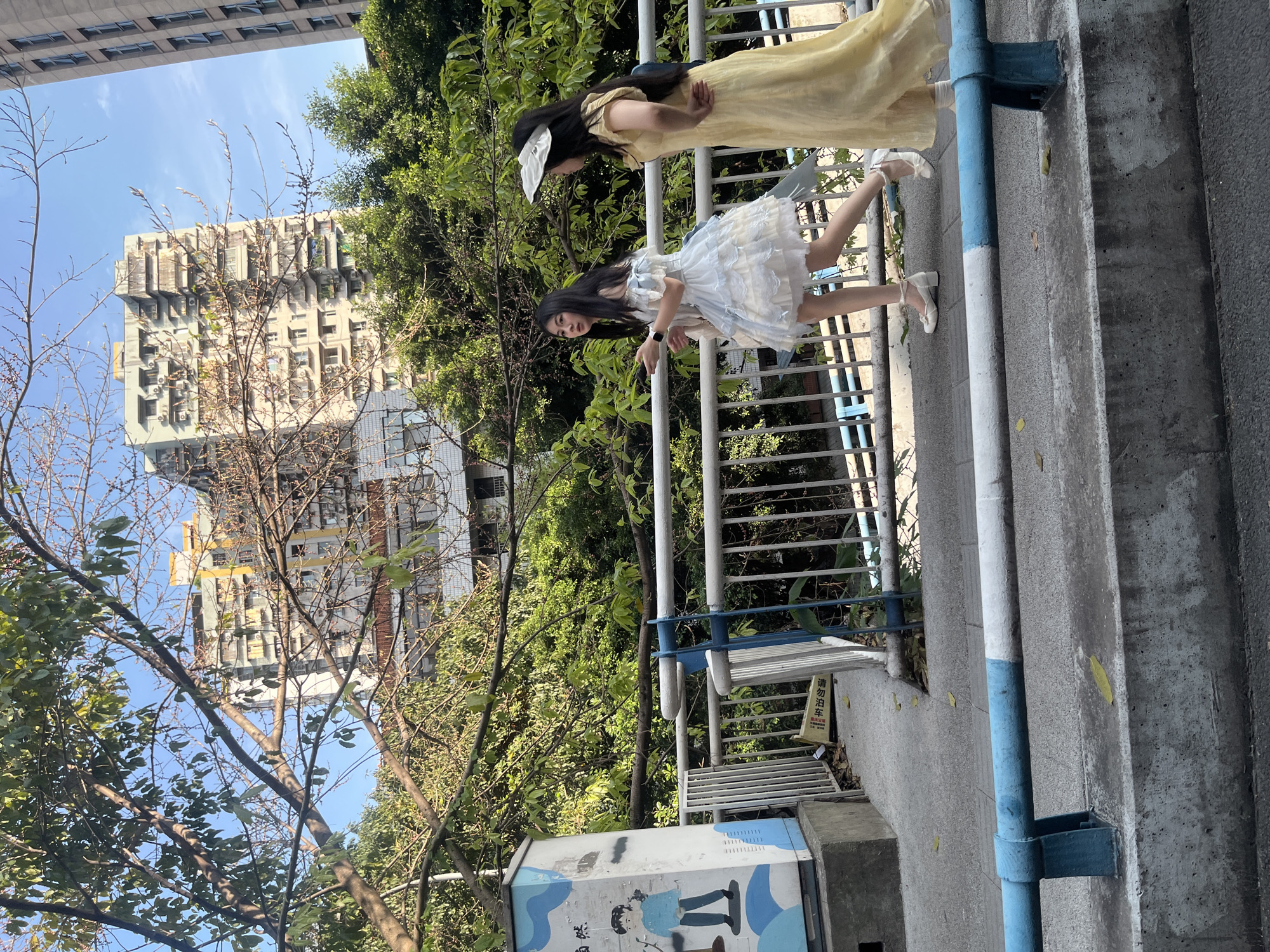 This media file captures an outdoor urban scene, presented with a 90-degree counter-clockwise rotation, featuring two young women on a pedestrian walkway. The time of day is bright and sunny, indicated by clear shadows and a vibrant blue sky with sparse clouds.
In the foreground, a woman with long dark hair, wearing a white or light blue ruffled short dress and white ballet-style shoes, is positioned next to a white and blue metal railing, looking towards the camera with a relaxed expression. Behind her, a second woman with long dark hair, adorned with a white bow, wears a long yellow dress and white shoes, standing with her hands clasped behind her back.
The walkway is concrete, with a white and blue painted pipe or support structure running along the side. A small white sign is attached to the railing, displaying Chinese characters that read "请勿泊车" (Qǐng wù bó chē), meaning "Please do not park." A utility box or pedestal with a light blue and white design, featuring cartoon figures, is partially visible at the bottom left.
The background is rich with greenery, including lush trees and some with bare or sparse branches. Two distinct buildings are visible: one with a unique, modern architectural style characterized by numerous protruding elements and what appear to be balconies or air conditioning units, and another taller, more conventional high-rise building. The scene depicts a casual moment in a well-developed urban environment, consistent with a location in Liangjiang, China.