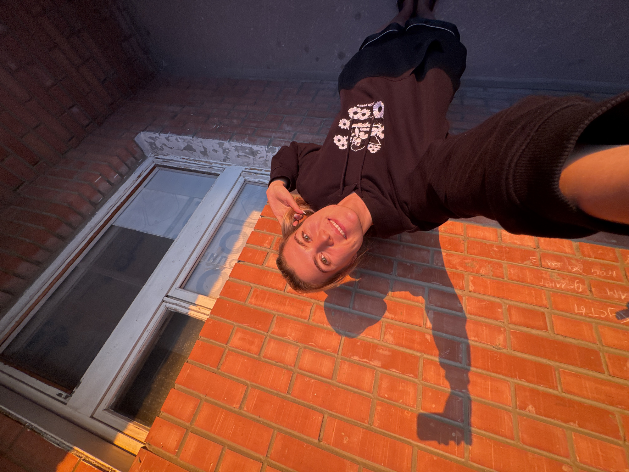 A young adult female is captured in a wide-angle, upward-looking selfie, taken outdoors next to a brick building in the city of Unknown, Unknown. She has light-colored hair, blue/green eyes, and a smiling expression, looking directly at the camera. She wears a dark (likely black) hooded sweatshirt adorned with a white graphic print featuring cherubic figures, daisy-like flowers, and the text "rest in peace." Below the sweatshirt, dark shorts are visible. Her right arm is extended, holding the camera, while her left hand is positioned near her left ear.
The background consists primarily of a reddish-orange brick wall with light-colored mortar. White Cyrillic script graffiti is present on the lower left section of the wall, with discernible words such as "усты" and "делают." To the right, a white-framed window with multiple panes is integrated into the brick wall. The ground beneath the woman is dark, suggesting concrete or pavement, partially in shadow.
The scene is illuminated by strong, warm, low-angle sunlight, indicative of either sunrise or sunset. This lighting casts a prominent, elongated shadow of the woman and her extended arm holding the camera onto the brick wall to her left. The shadow clearly delineates the shape of her head, arm, and the rectangular device (presumably a smartphone) in her hand.
