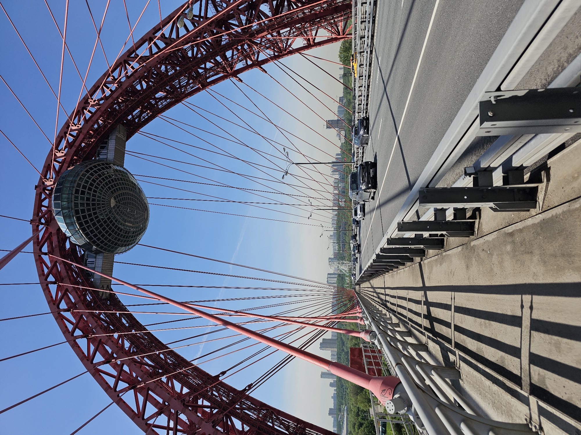 The image shows a view from the Zhivopisny Bridge in Khimki, Russia. The bridge is a cable-stayed bridge spanning the Moskva River. The focal point of the image is the large red arch of the bridge and the spherical structure suspended within the arch. The structure is clad in a grid-like pattern. Steel cables extend from the arch downwards towards the roadway. Traffic is visible on the bridge's roadway, with several cars traveling along the asphalt surface. A concrete barrier separates the roadway from the pedestrian or maintenance area. The environment includes blue skies overhead, with a slight haze in the distance where city buildings and greenery are visible below the bridge. Shadows are cast by the railings. The image appears to have been taken from a low angle, looking upwards and outwards across the bridge.