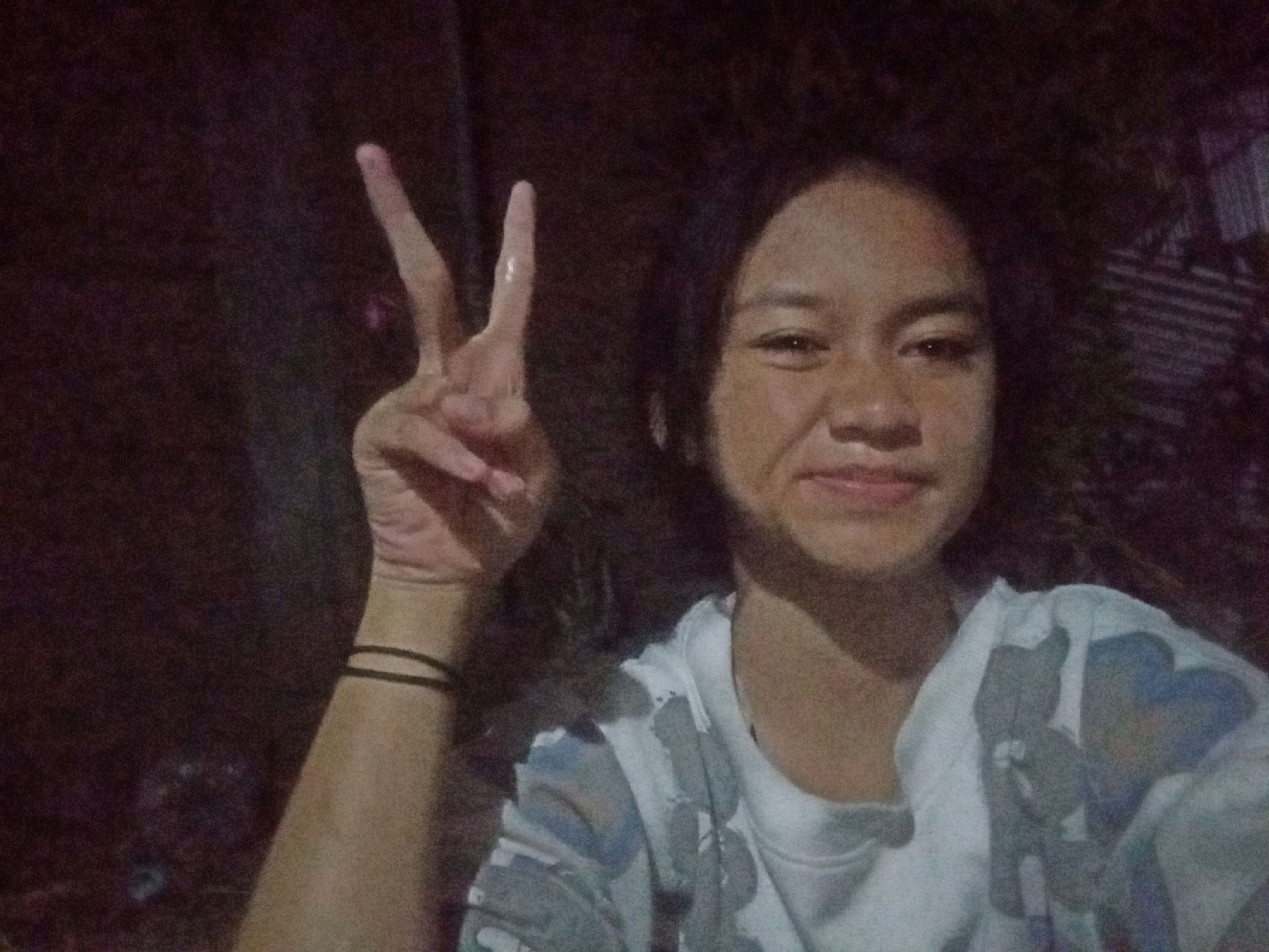 A young person with dark, shoulder-length hair is taking a selfie at night. They are smiling and holding up two fingers in a peace sign. They are wearing a white t-shirt with a colorful, abstract pattern of grey, blue, and pink shapes, possibly depicting cartoon characters. A black wristband is visible on their left arm. The background is dark and appears to be outdoors, with hints of vegetation and a corrugated metal fence or structure on the right side. The lighting is dim, suggesting it is nighttime. The image quality is grainy, indicating it may have been taken with a low-light setting on a mobile device. The location is identified as Badas, Indonesia.