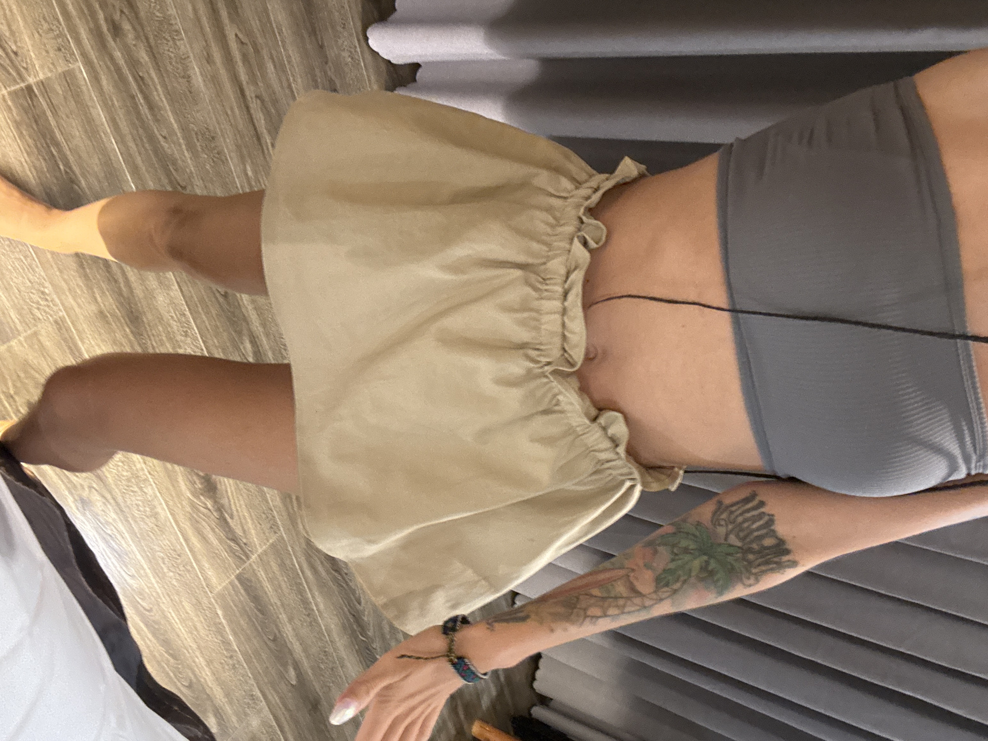 In a domestic setting, likely a bedroom, a person is standing in front of a grey curtain. They are wearing a grey strapless top and a tan ruffled mini-skirt. The person has tattoos on their right arm and is wearing a bracelet. The flooring is made of wooden planks. The image does not provide enough information to determine the specific location within Nam Hoa Lư Ward, Vietnam.