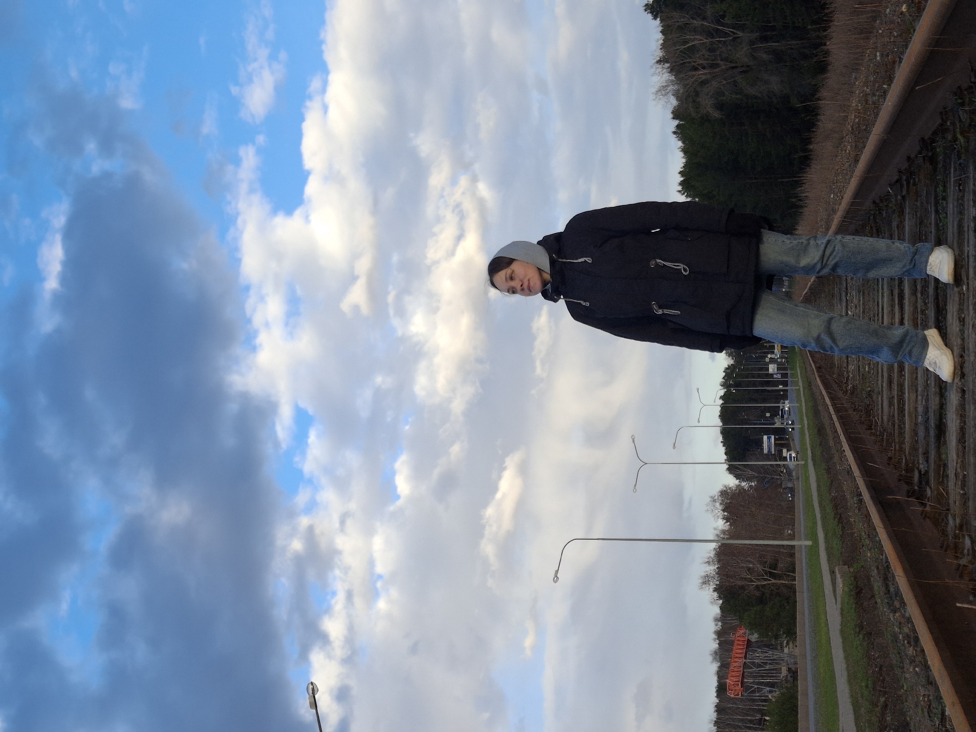 The image captures a full-body shot of a person standing on railway tracks under a partially cloudy sky. The perspective is rotated 90 degrees clockwise, making the person appear to be standing vertically on the tracks.
The main subject is a young adult, wearing a dark (likely black) hooded jacket with distinctive toggle closures, light blue jeans, and white sneakers. The jacket's hood is up, framing their face, which has a neutral and slightly pensive expression as they look downwards and to the side.
The setting is outdoors, alongside a set of railway tracks that extend into the distance. The tracks are rusty, with visible wooden sleepers. To the right of the tracks (when viewing the image upright), there is a strip of bare earth and then a dense line of evergreen trees. To the left, parallel to the tracks, runs a paved path or sidewalk, and beyond that, a road with a line of modern streetlights. Distant vehicles are visible on the road, and more trees, some bare, line the far side of the road. In the far distance on the left, a large orange construction crane is partially visible.
The sky is a prominent feature, displaying a mix of bright blue patches and various cloud formations, including fluffy white cumulus clouds and darker, heavier grey clouds, suggesting dynamic weather or a time around sunrise/sunset. The overall lighting is natural and clear, indicating daytime. No legible text is visible on any signs or objects within the frame. The scene conveys a sense of quiet observation in a semi-urban or industrial-adjacent environment.