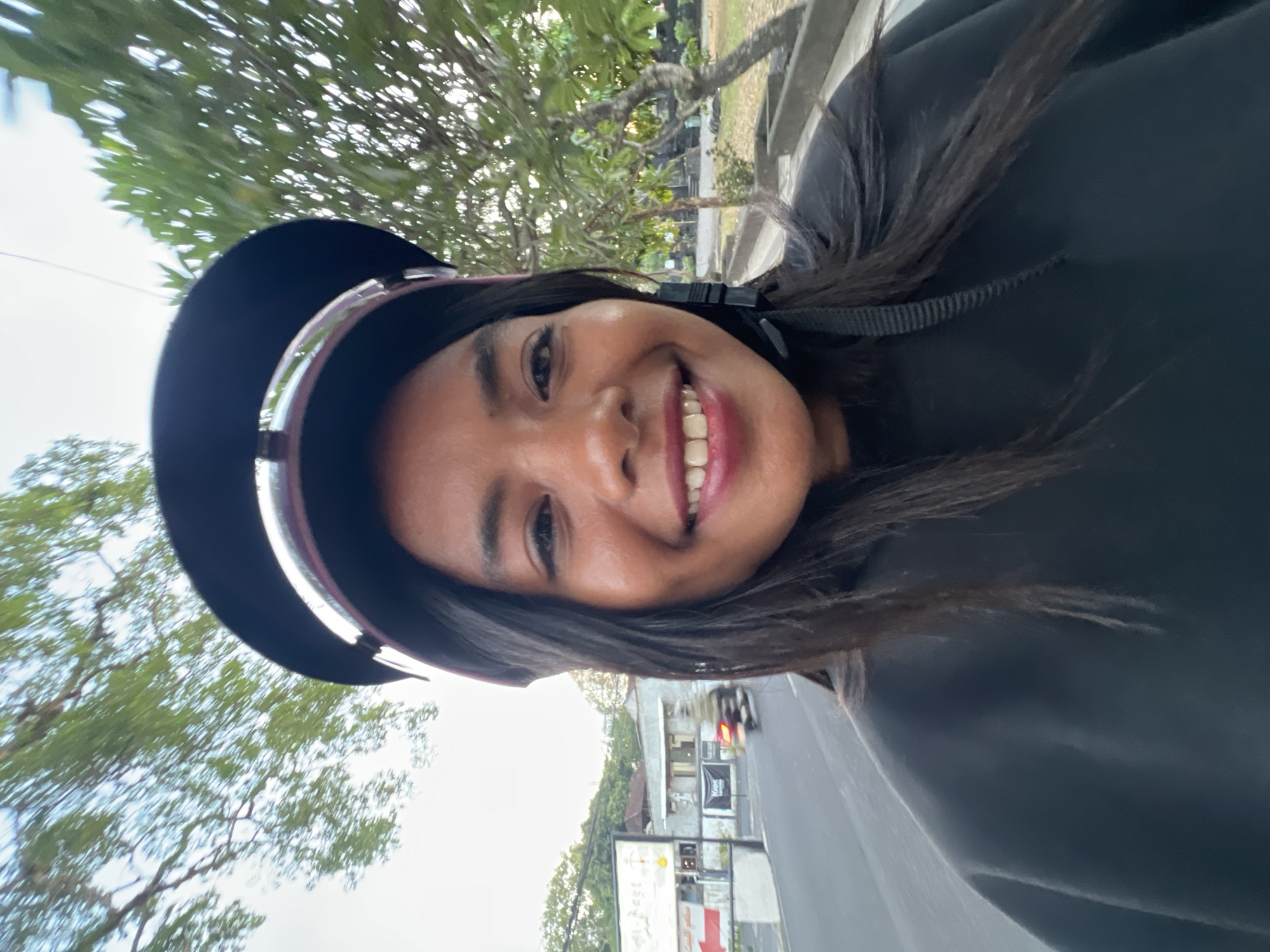A woman with dark hair and a helmet is taking a selfie while riding a motorcycle in Kuta, Indonesia. The woman is wearing a black sweatshirt and her face is framed by the helmet. She is smiling at the camera. In the background, there are trees and buildings. The road appears to be busy with traffic.