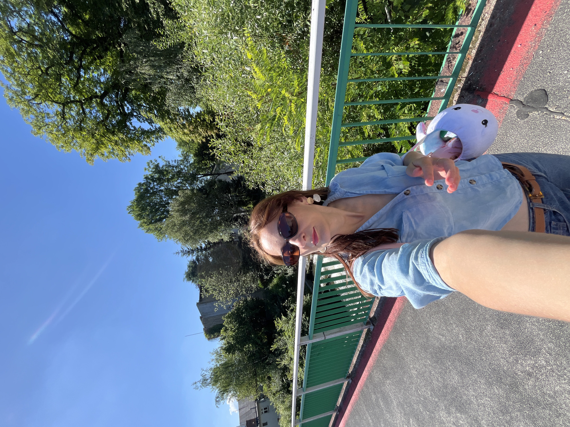 A woman with long, brown hair and sunglasses takes a selfie on a bridge. She is wearing a light blue denim shirt and blue jeans. In her right hand, she holds a stuffed white bunny bag. The bridge has a green metal railing with a white top rail and a red border on the walkway. Behind her, lush green trees and buildings are visible under a clear blue sky. The bridge appears to span over a body of water, suggested by a glimpse of moving water below the railing.