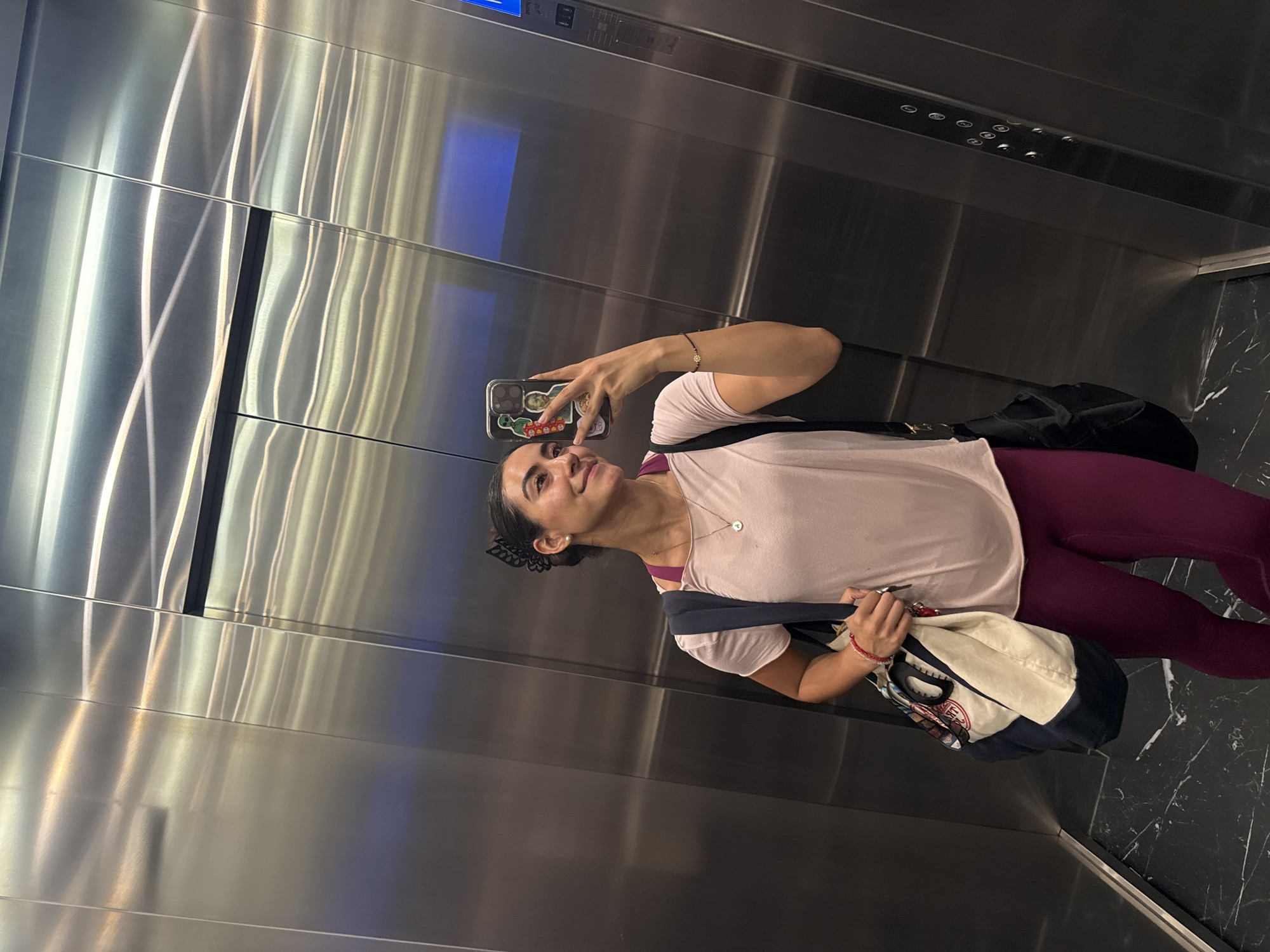 A woman is taking a selfie in an elevator. She is wearing a light pink t-shirt and maroon leggings. She has two bags slung over her shoulder, one black and one cream and navy. The elevator interior is stainless steel with a black marble floor. The elevator's control panel is visible in the background. The location appears to be in Puebla, Mexico.