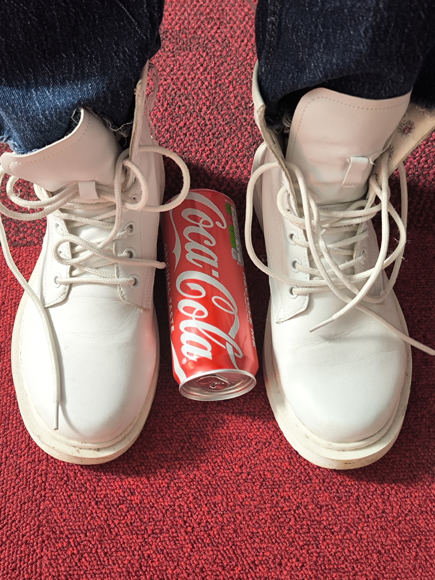 The image shows a Coca-Cola can positioned between two white boots. The boots are worn by someone wearing blue jeans. The background is a red carpet. The location and city are not identifiable from the image.