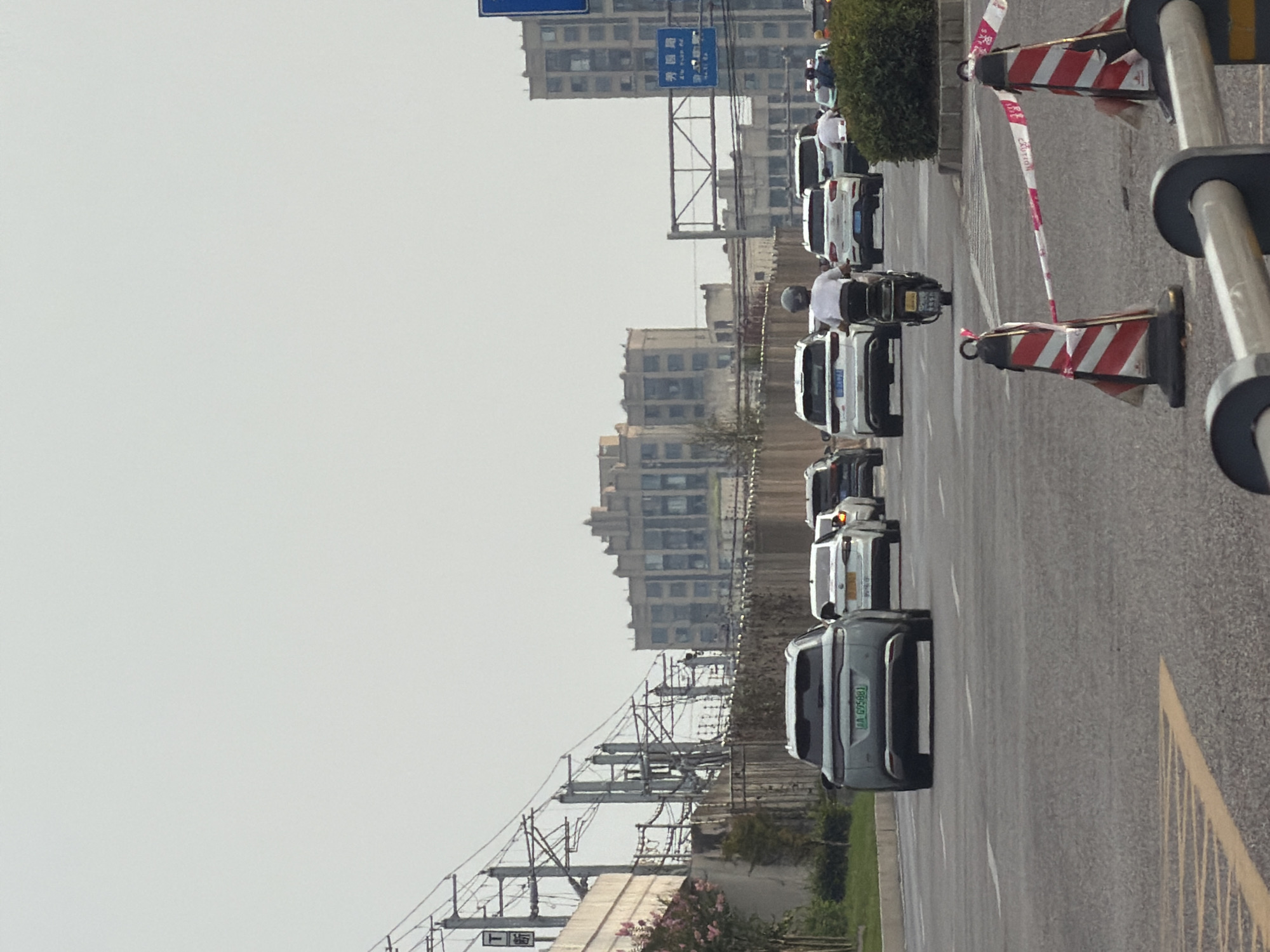 The image captures a view of a traffic scene in the city of Jiangjin, China, under a hazy sky. In the foreground, traffic cones with red and white striped caution tape cordon off an area of the asphalt road. Several vehicles are proceeding forward, including a light blue SUV with a green license plate in the left lane, followed by a white car. In the right lane, a person on a scooter is riding ahead of other vehicles, including two white SUVs and another car. In the background, a series of utility poles with extensive wiring dominate the left side of the frame, leading to several multi-story buildings. A prominent, larger building with a distinctive stepped facade is visible further back, and another tall building is situated on the right. Greenery lines the side of the road, and bushes are visible on the far right. Blue street signs are mounted on poles.