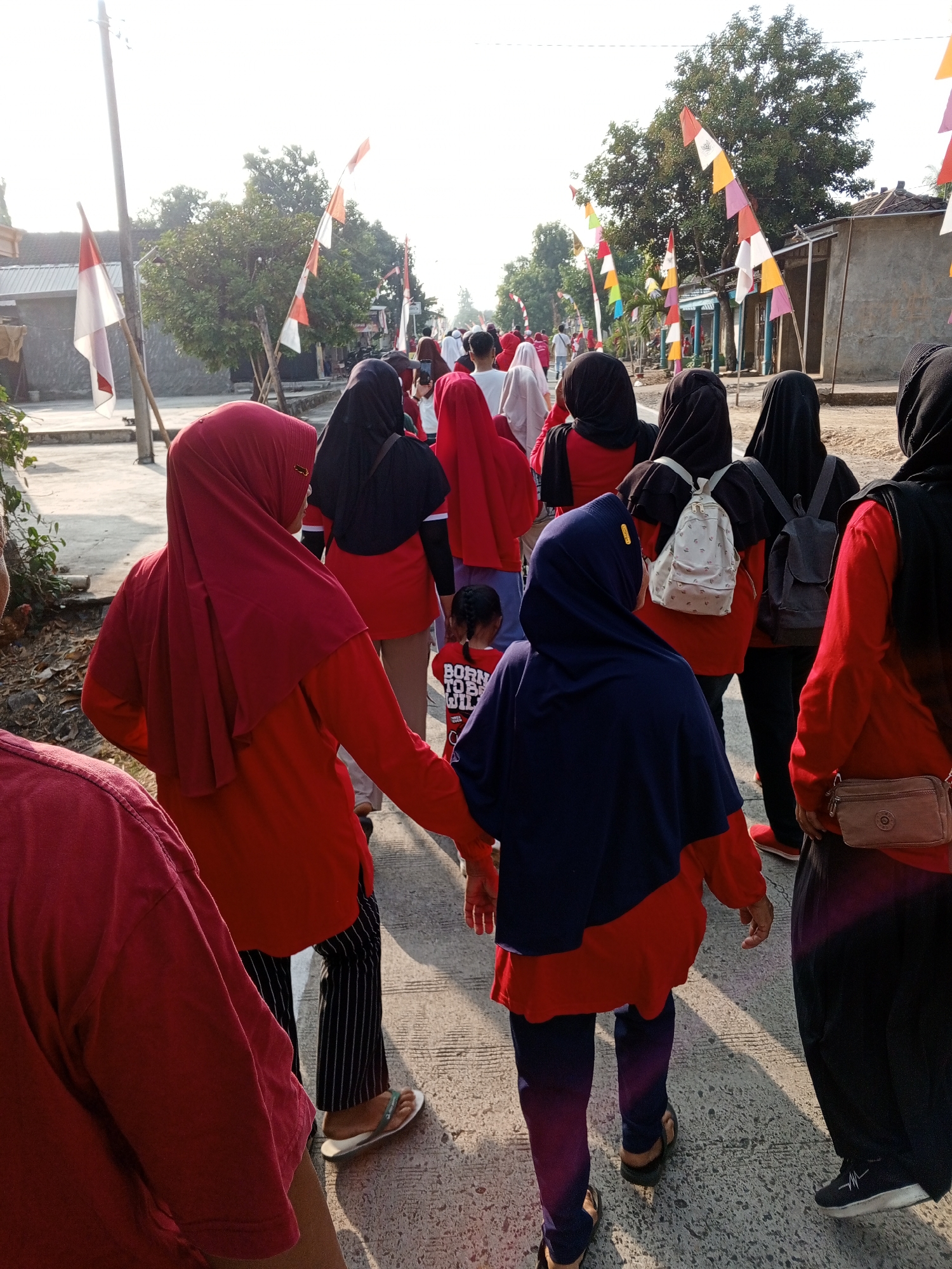 In Lemahbang, Indonesia, a group of people, mostly women wearing hijabs and red tops, are walking along a paved road. Some are carrying backpacks, and one person is wearing striped pants and flip-flops. The road is lined with buildings, trees, and flags. The sky is bright and sunny. The scene suggests a cultural or religious event is taking place.
