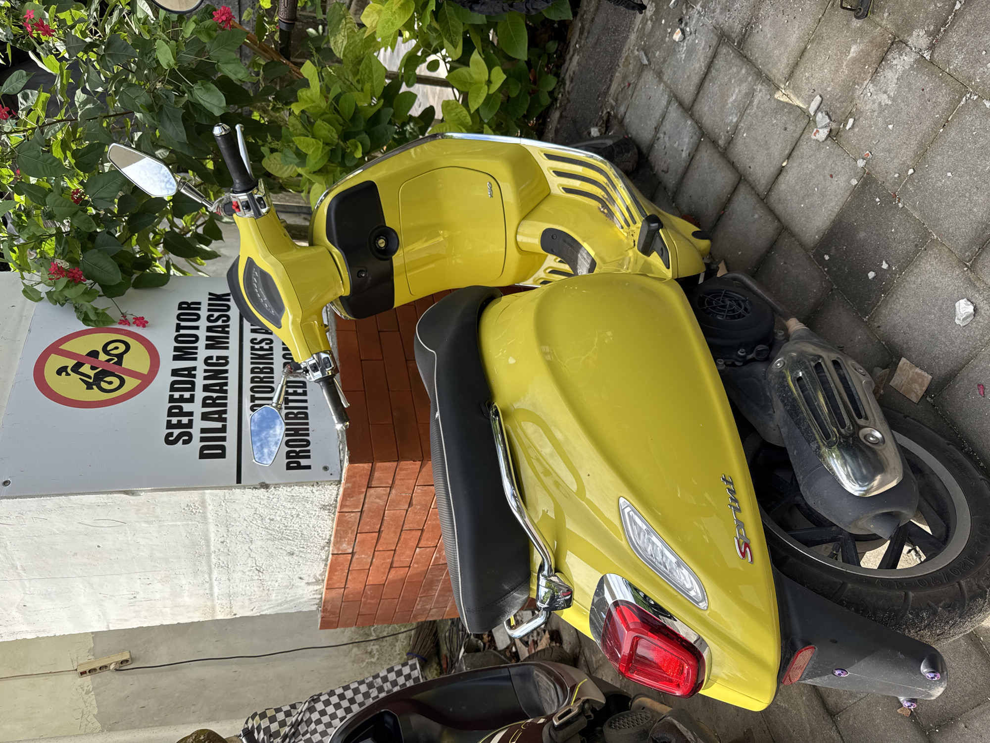 A bright yellow Vespa scooter is parked on a brick sidewalk next to a sign reading "SEPEDA MOTOR DILARANG MASUK MOTORBIKES PROHIBITED," presumably in Kuta Selatan, Indonesia, given the language on the sign. The scooter is parked perpendicular to the sign and the brick building behind it. The scene is outdoors, with foliage visible behind the sign. Another vehicle, partially obscured, is visible in the lower left of the frame. The environment appears to be urban, with paved surfaces and buildings.