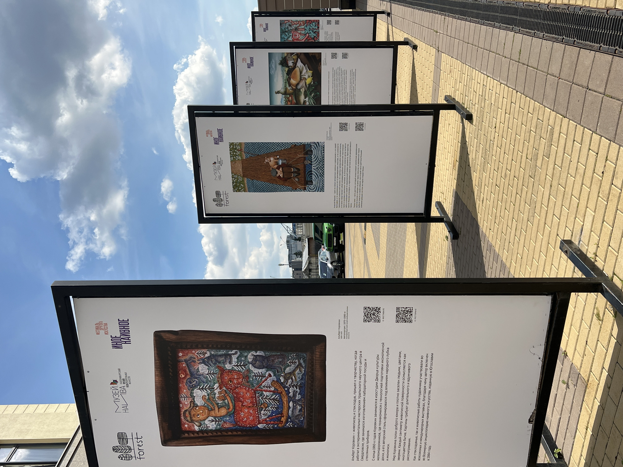 The image depicts an outdoor exhibition of naive art in Yekaterinburg, Russia. Three large, freestanding display panels are set up on a paved walkway, likely in a public square or park. The panels feature reproductions of paintings with descriptive text and QR codes. The first panel, on the left, prominently displays a painting by Albert Korovin, a well-known naive artist from Yekaterinburg. The painting depicts a fantastical scene with a human-like figure, a red bull-like creature, and various animals in a forest setting. The text on the panel provides biographical information about Korovin and describes his artistic style. The second panel displays another artwork, and the third panel shows a still life with fish and fruit. The exhibition is part of a festival organized by the Museum of Naive Art in Yekaterinburg, as indicated by the logos and text on the panels. The background shows a building, a street with cars, and a blue sky with scattered clouds. The overall scene suggests a cultural event taking place in a contemporary urban environment.