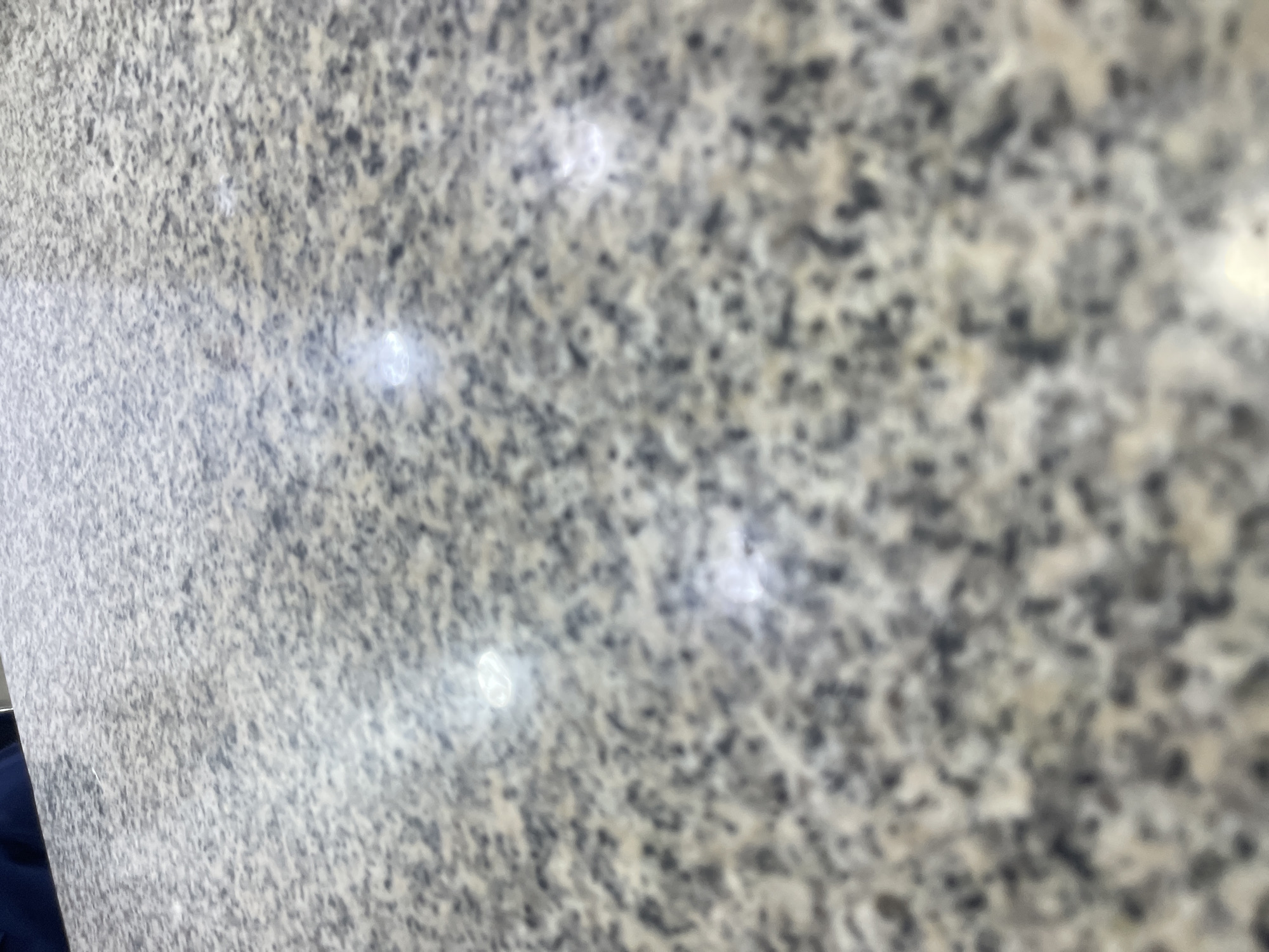 The image shows a close-up, slightly out-of-focus view of a speckled granite countertop. The pattern of the granite consists of small flecks of black, gray, and off-white. Several bright, blurry spots on the surface suggest reflections from overhead lighting. In the upper left corner, a small portion of dark blue fabric is visible, likely clothing. The perspective is from slightly above and to the side of the countertop, looking down and away from the viewer. The scene appears to be indoors, possibly in a kitchen or a commercial establishment like a restaurant or shop. The granular texture of the granite is the primary focus. There is no indication of the specific location in Jos, Nigeria, beyond the assumption that this is a photograph taken within that city. The image is devoid of any context that would place it geographically.