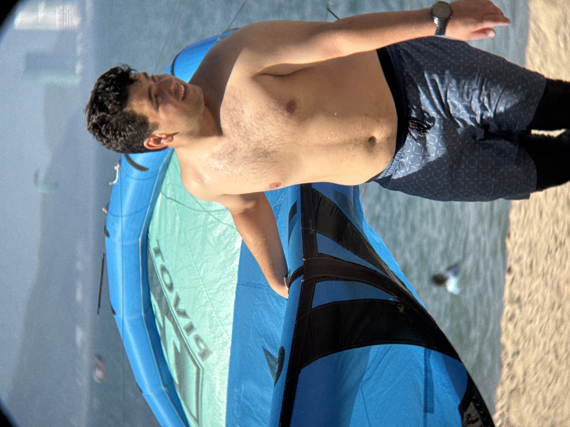 An image depicts a young man, shirtless and wearing patterned swim shorts, standing on a sandy beach. He is holding a large, blue kite. The kite has some text on it, which appears to read "PIVOT". In the background, the ocean is visible, with the blurry shapes of other kitesurfers in the distance and a hint of buildings on the horizon, suggesting the urban coastline of Nha Trang, Vietnam. The man is looking to his right and smiling.