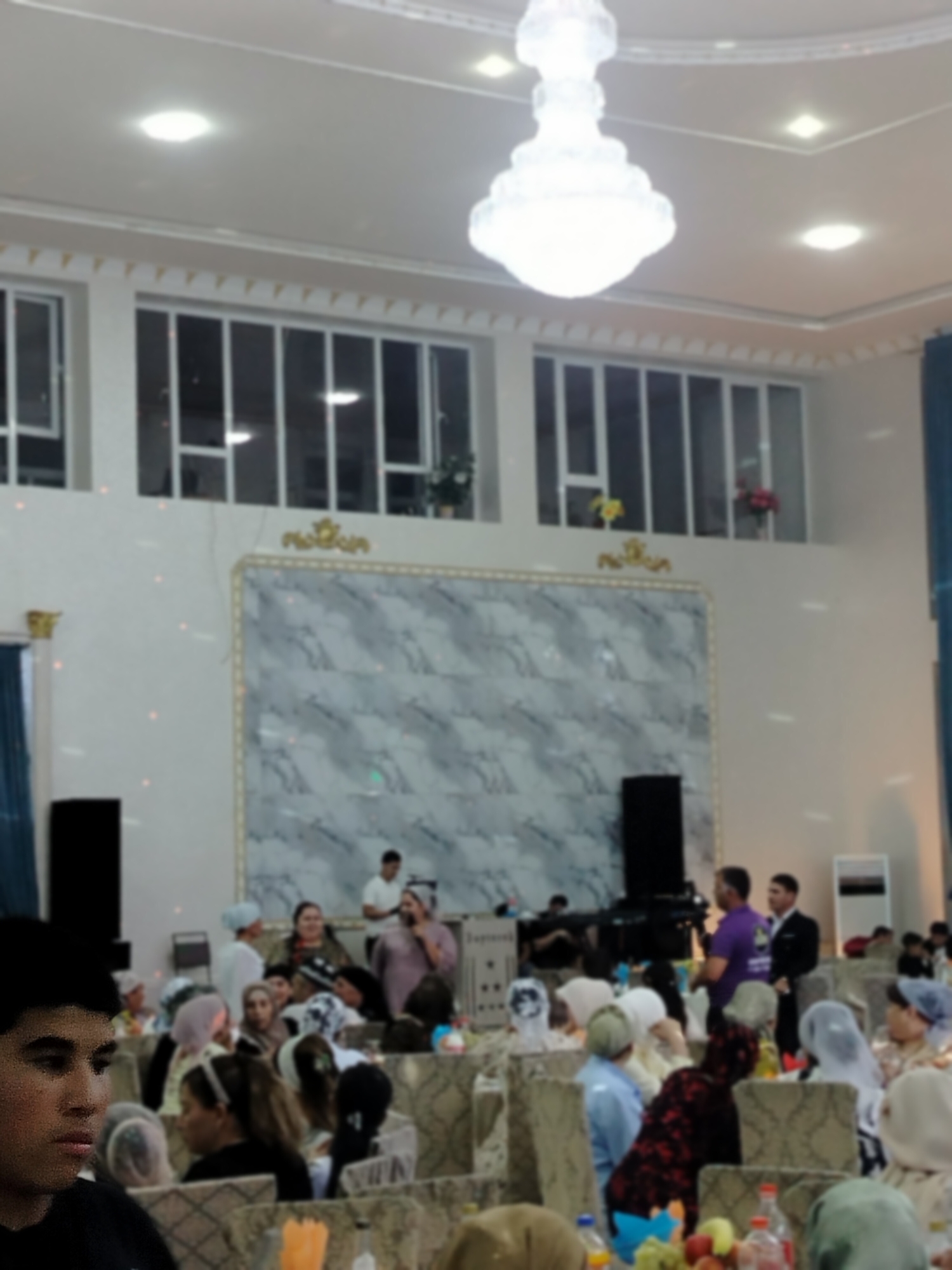 The image depicts an indoor gathering, likely a celebration, in Ақсуабат, Kazakhstan. A large chandelier hangs from the ceiling, illuminating the room. The walls are decorated with a large marble-patterned panel and gold accents. Several tables are set for dining, with people seated and interacting. Some individuals are standing, including a man speaking into a microphone. The overall atmosphere appears to be festive.