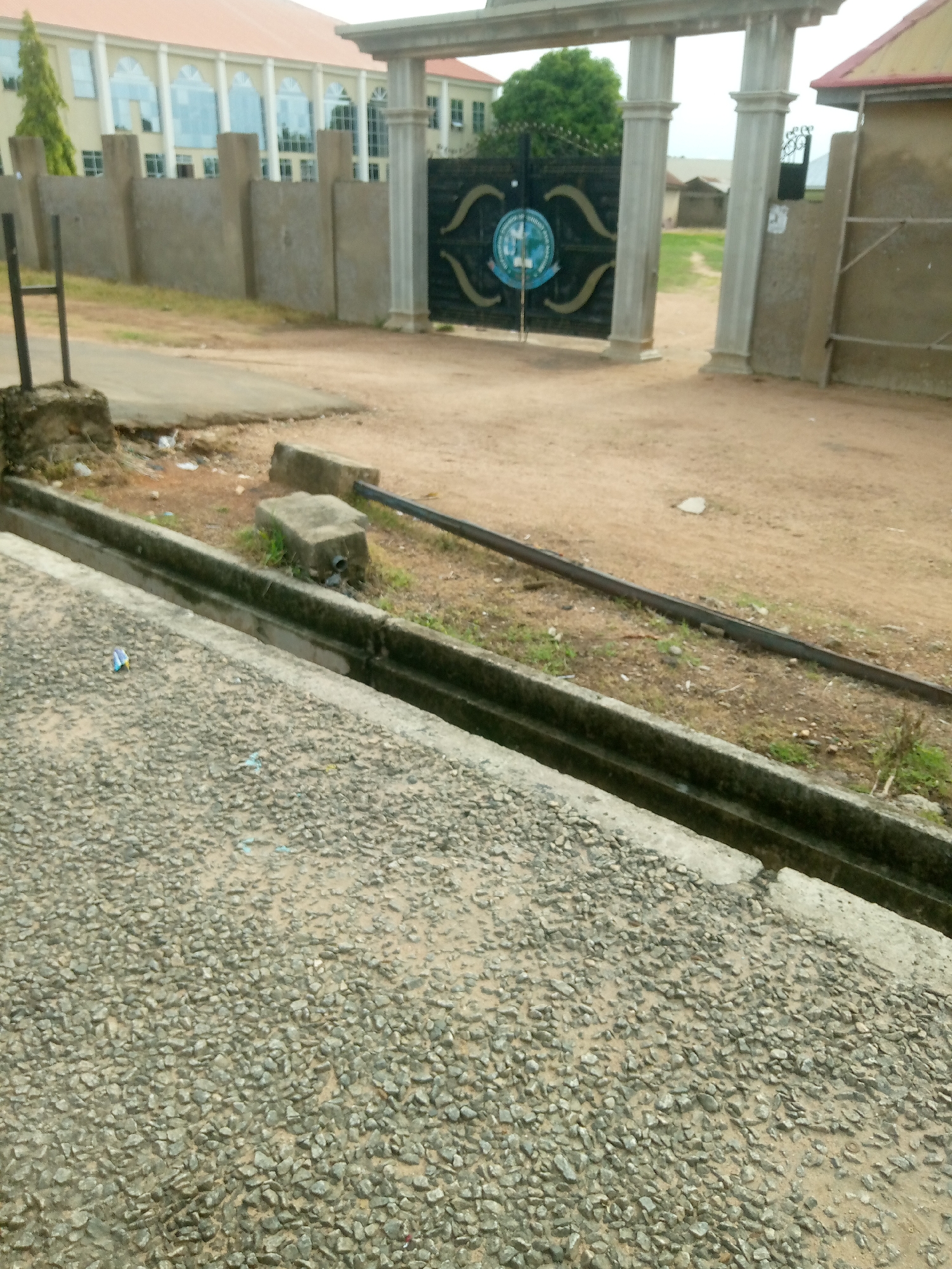 The image depicts the entrance to a compound in Jalingo, Nigeria. A large, ornate black gate with a circular blue and white emblem is visible, flanked by two substantial stone pillars. Beyond the gate, a dirt path leads into the compound, with glimpses of buildings and trees. A low concrete wall runs along the side of the compound, and in the foreground, there is a gravel-covered surface with a drainage ditch. Scattered debris, including a small blue and white wrapper, is present in the ditch and on the gravel. The overall scene suggests a public or institutional entrance in a tropical climate, with evidence of wear and tear on the structures and surrounding environment.