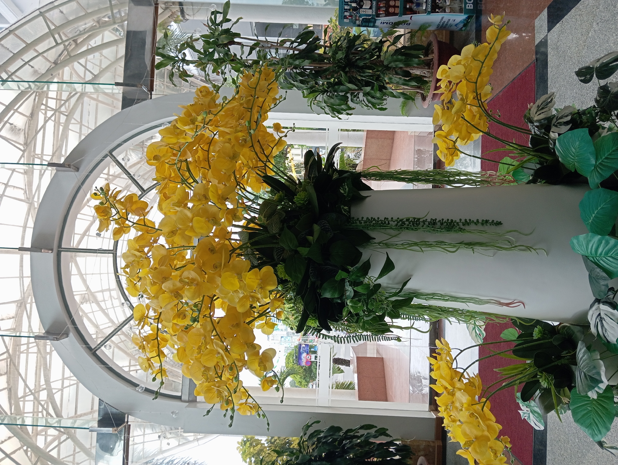 In Nha Trang, Vietnam, an indoor scene features a large, white, cylindrical vase prominently displaying an abundant arrangement of artificial yellow orchids. The orchids, in various stages of bloom, cascade from the top of the vase, their stems arching gracefully. Surrounding the base of the orchids are dark green leaves and several trailing, lighter green vine-like plants. To the right of the main floral arrangement, a tall potted plant with dark green, elongated leaves stands. Behind the flowers, a large arched window with a translucent, grid-like ceiling structure suggests an atrium or conservatory setting. Through the window, a glimpse of an outdoor environment is visible, including palm trees and architectural elements of buildings. In the foreground, the floor is a tiled mosaic of grey and white squares, with a red rug partially visible. To the far right, a shelf unit displays various bottles and packaging, suggesting a retail or service area. No people are visible in the image.