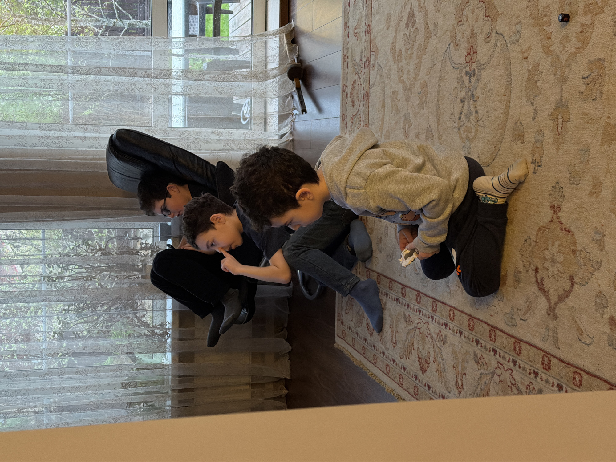 The image, taken at an elevated angle, depicts three children indoors on a patterned rug. One child, wearing a gray hoodie and mismatched socks, kneels and holds a small white object. Another child points at their nose. A third child in glasses leans forward. A black object, possibly a guitar case, rests near a window with patterned blinds. A small black object is visible on the rug. The location is Новоглаголево, Russia.