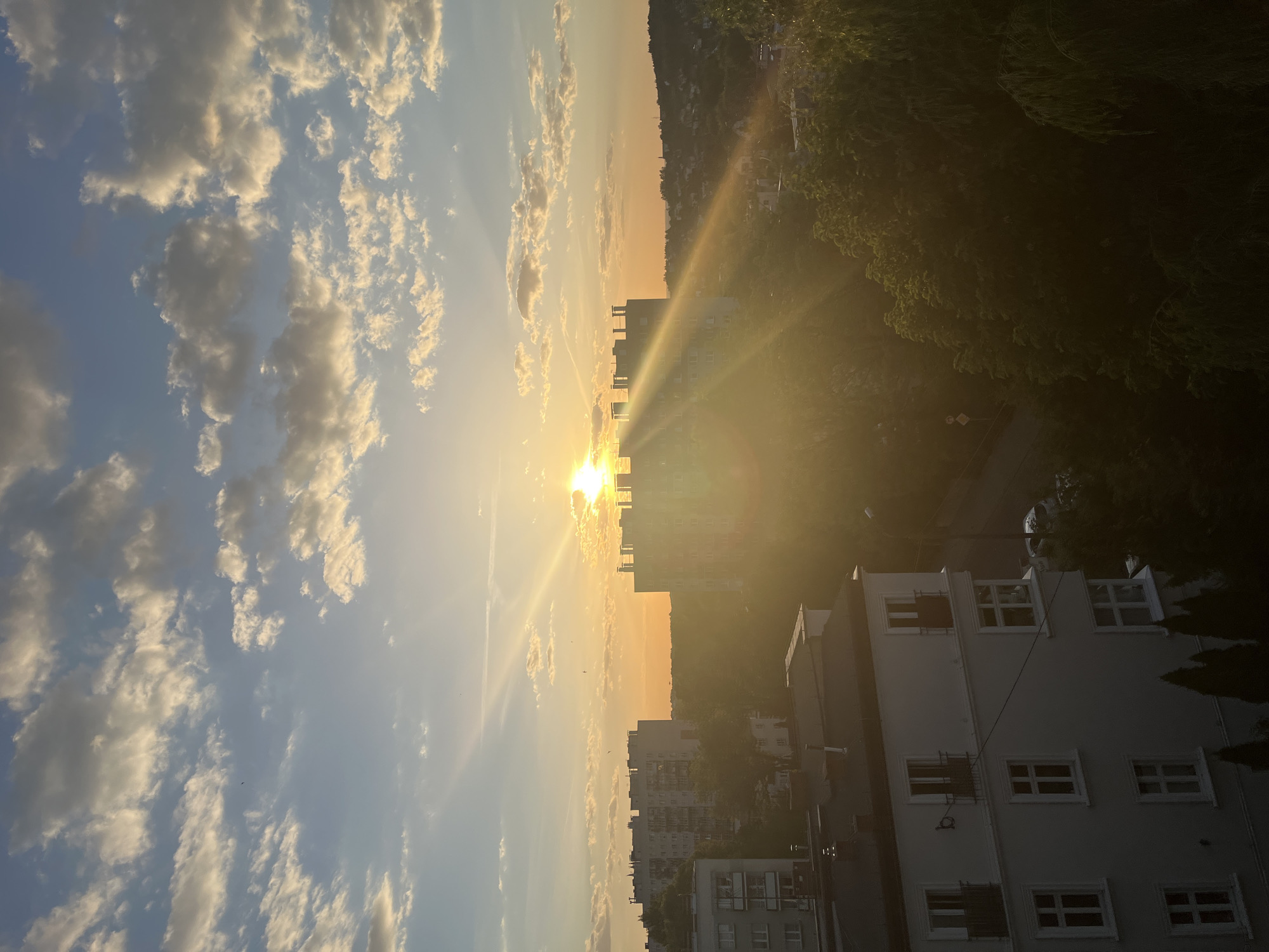 The image depicts a sunrise in a city. The sun is low on the horizon, casting a bright glow and long rays across the scene. The sky is filled with clouds, some reflecting the golden light of the sunrise. Below the horizon, there are apartment buildings of various heights, silhouetted against the bright sky. To the right of the buildings, there is a dense area of trees and vegetation. In the lower right corner, a multi-story building with numerous windows is visible. The location is Unknown, Unknown.