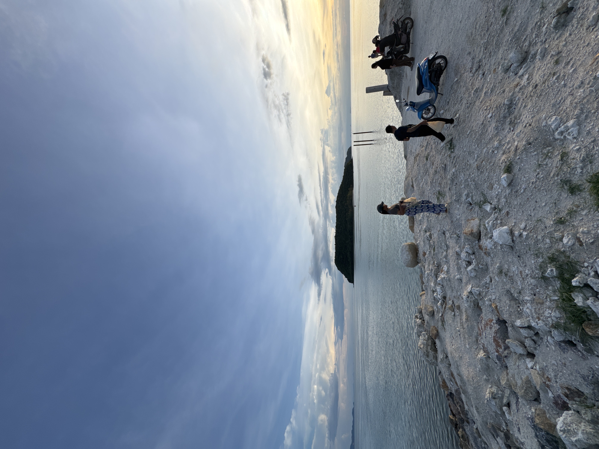 A wide-angle, eye-level outdoor shot captures a serene coastal scene at sunset on Koh Phangan, Thailand. The foreground features a rocky, uneven shoreline with sparse green vegetation. Several people are present, interacting with a blue scooter and two motorcycles parked on a sandy, slightly elevated area. To the left, a woman in a cap and patterned pants stands holding a white bag. To her right, a man in dark clothing walks with a white bag. Further right, two individuals are near the motorcycles, with one person appearing to be on a motorcycle. In the middle ground, the calm, blue water stretches out to the horizon, meeting a sky painted with soft clouds and the warm hues of the setting sun. A small, tree-covered island is visible in the distance, silhouetted against the sky. Three wooden posts rise from the water in the mid-distance. The overall atmosphere is peaceful and contemplative.
