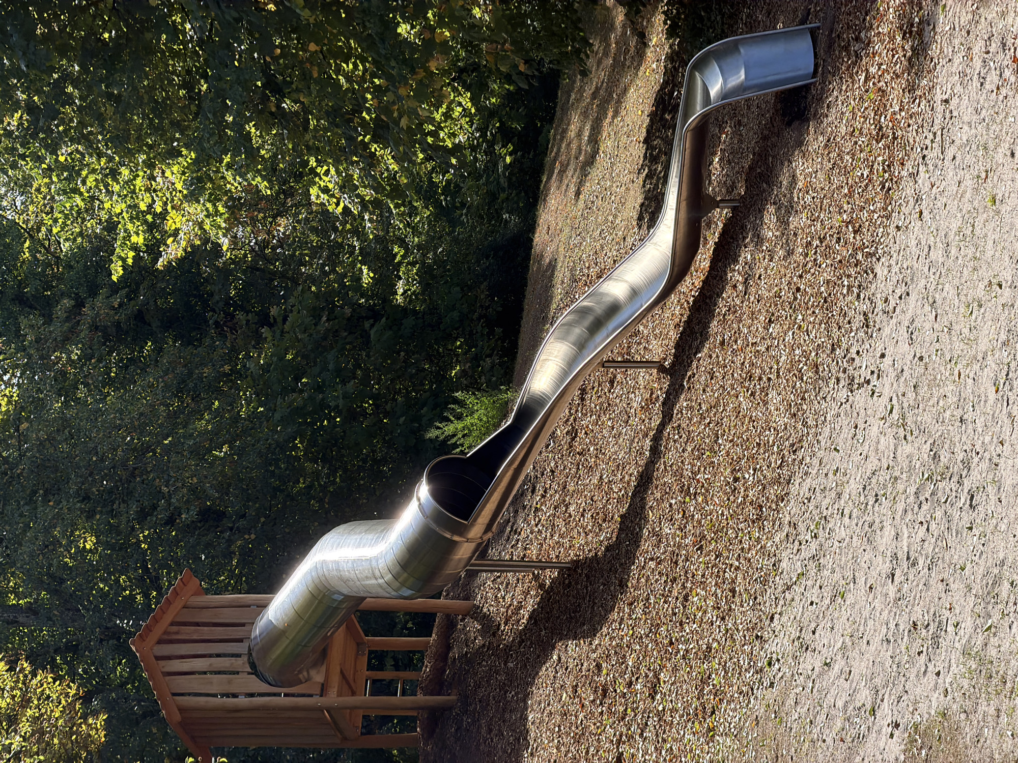 A stainless steel tube slide is situated in a wooded area, likely a park or playground in Esch-sur-Alzette, Luxembourg. The slide emerges from a wooden playhouse structure elevated on stilts. The ground beneath the slide is covered with a layer of wood chips and fallen leaves, transitioning to sandy soil with sparse grass in the foreground. Sunlight dapples through the dense green foliage of mature trees that form the backdrop and canopy of the scene, casting shadows on the slide and ground. No people are visible in the image.