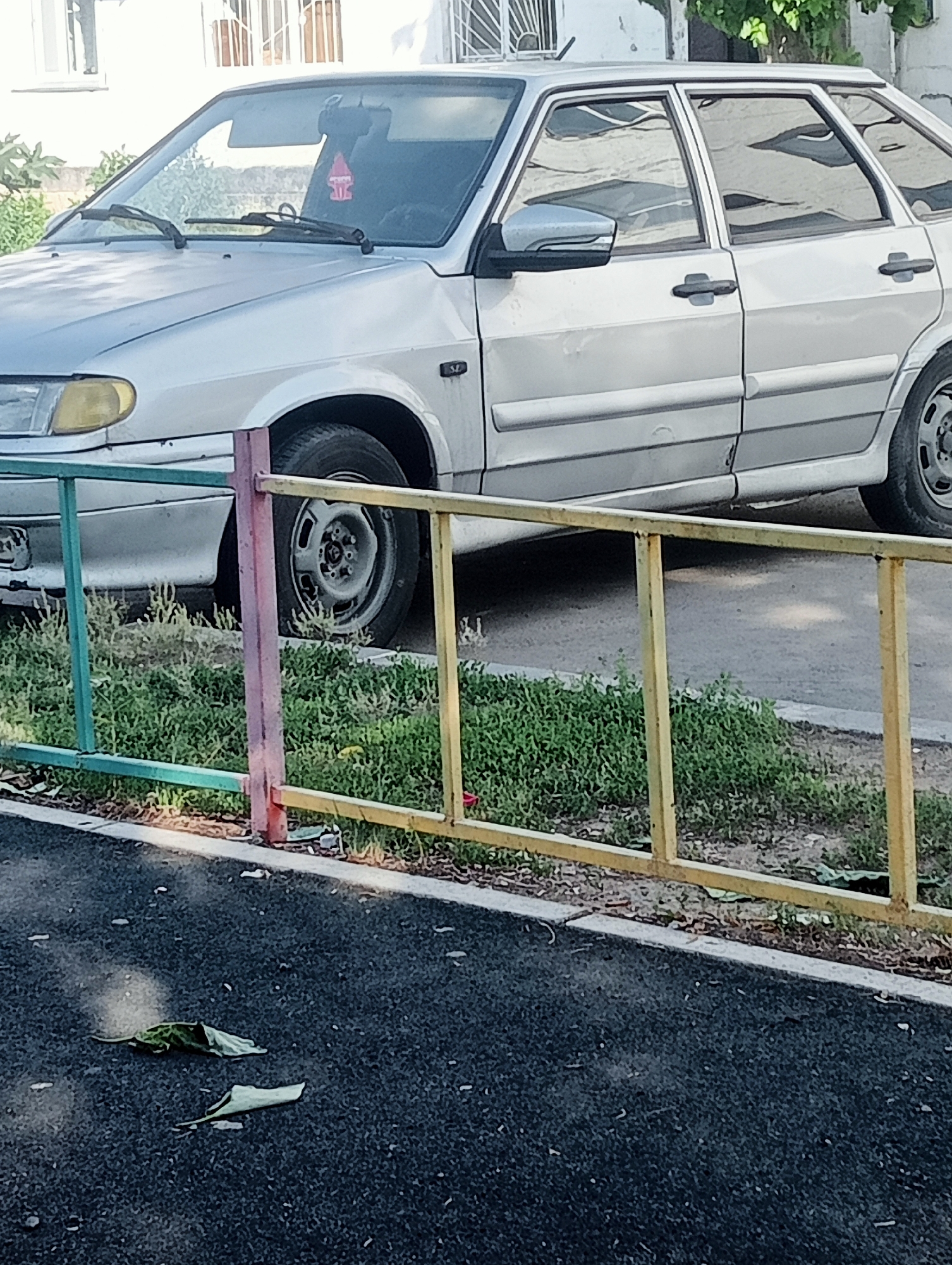 The image shows a silver hatchback car parked on a street. The car is partially obscured by a multi-colored metal fence. The street appears to be in a residential area, with a building visible in the background. There are some scattered leaves on the ground. The car's right side is facing the camera, and the front right wheel is visible. The car has its driver's side door open. There is a air freshener hanging from the rearview mirror.