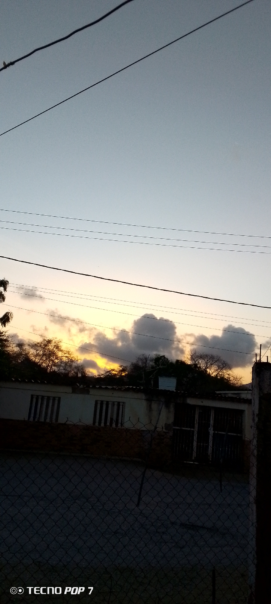 The image shows a scene in Altagracia, Venezuela, taken at dusk or dawn. The sky is a gradient of light blue to yellow, with a line of darker clouds along the horizon. Three electrical lines run horizontally across the top portion of the frame. Below, a partially obscured building is visible, with barred windows. A chain-link fence dominates the foreground. The lower left corner of the image displays the "TECNO POP 7" watermark.
