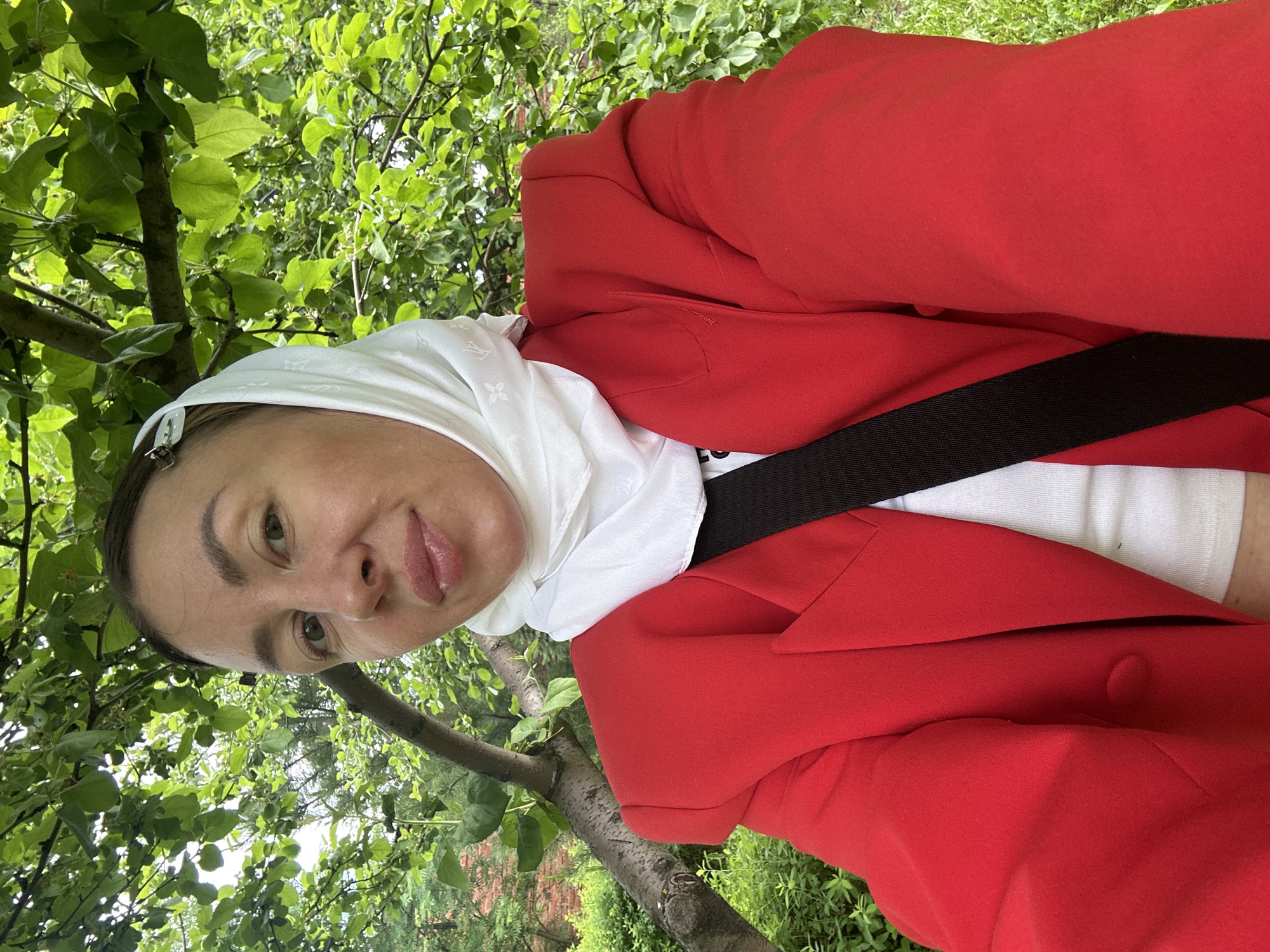 A woman is positioned in an outdoor environment with dense foliage, possibly within the city of Votkinsk, Russia. She is wearing a red blazer over a white top and a white headscarf with a pattern. A black strap, possibly from a bag, crosses her torso. The surrounding environment includes trees and greenery, with a tree branch visible to the left of the woman's face.