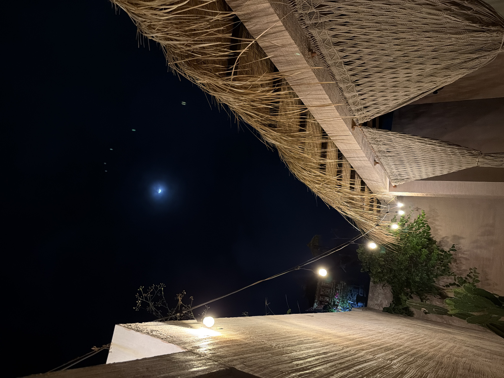 The image depicts a night scene in North Kuta, Indonesia, featuring a structure with a thatched roof and woven net-like decorations. A crescent moon is visible in the dark sky. String lights are strung along the structure. Plants, including a leafy bush and cacti, are growing nearby. A lit path extends into the background, where a dark opening or entrance is suggested.