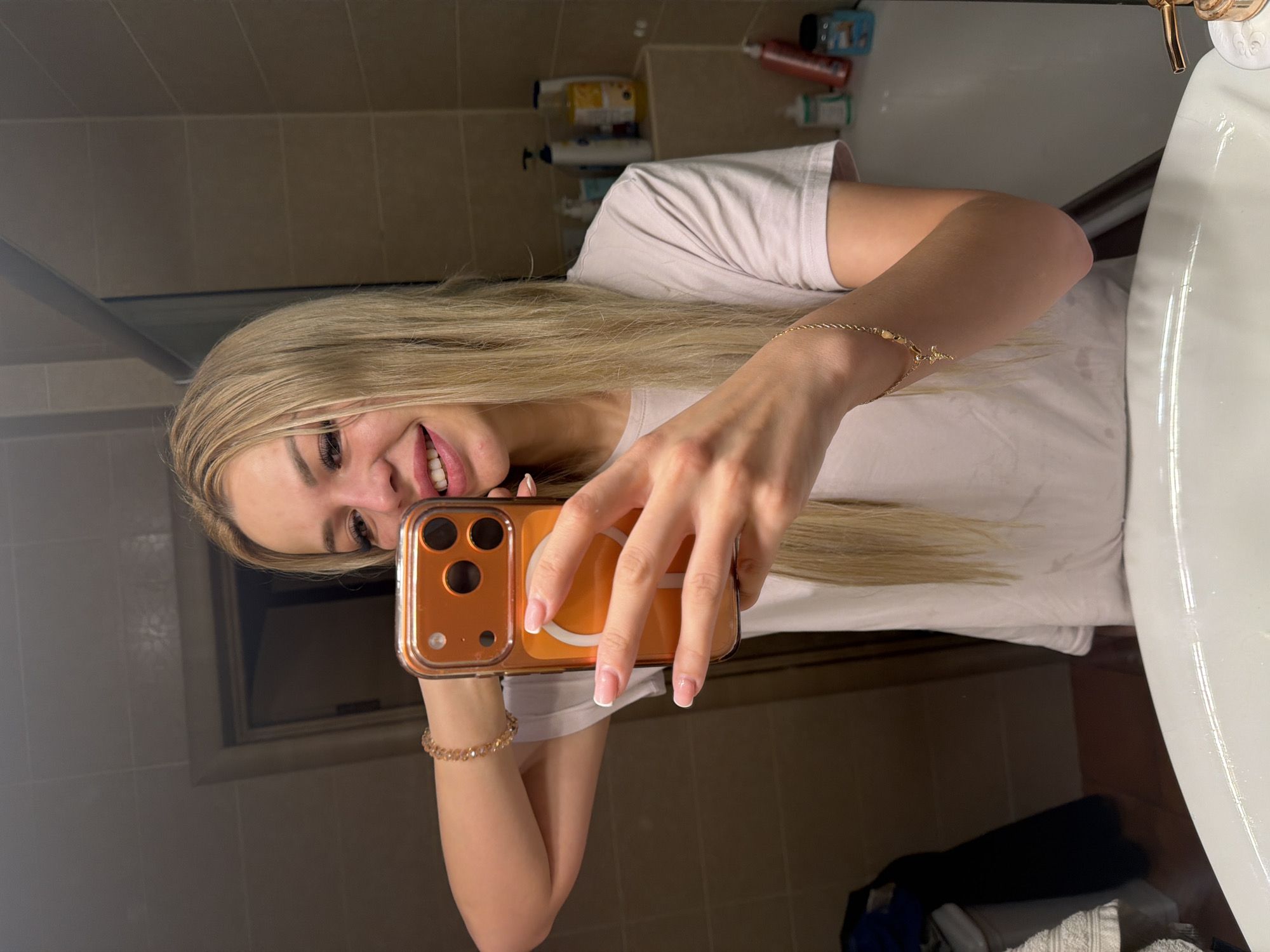 A young woman with long blonde hair and French manicured nails is taking a selfie in a bathroom. She is wearing a light-colored t-shirt and a gold bracelet with a cross charm on her left wrist, and another gold bracelet on her right wrist. She is holding an orange-cased smartphone in her hands. The bathroom has tiled walls and various toiletries on shelves in the background. The scene is lit by artificial light. The location is identified as a bathroom in Kyiv, Ukraine.