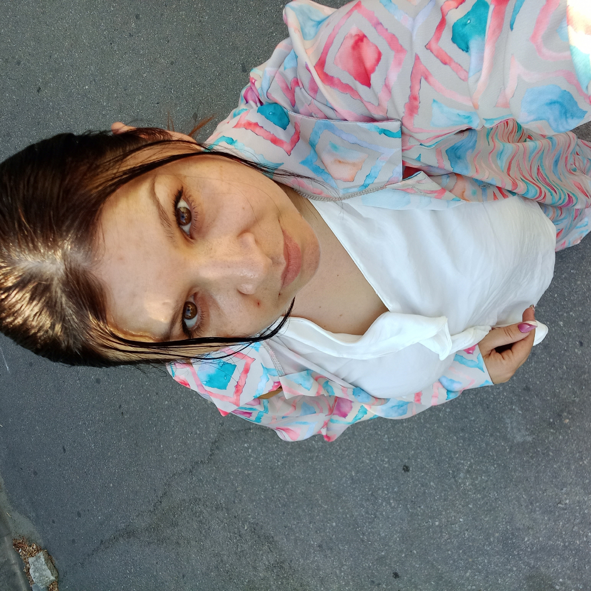 The image shows a woman from a high-angle perspective, standing on an asphalt surface. She has brown hair and brown eyes, and is wearing a white top and a light-colored jacket with a geometric pattern of pink, blue, and gray. Her right hand is visible, with painted fingernails. The asphalt surface appears to be a road or sidewalk, with some debris visible on the left side of the frame, including a concrete block and dry leaves. The lighting suggests it is daytime, with sunlight illuminating the woman's hair and face. There are no other people or objects of significance in the immediate surroundings. The overall impression is a candid street-style photograph taken outdoors in an urban environment. Based on the visual cues and common urban textures, this scene could be located in Kropyvnytskyi, Ukraine.