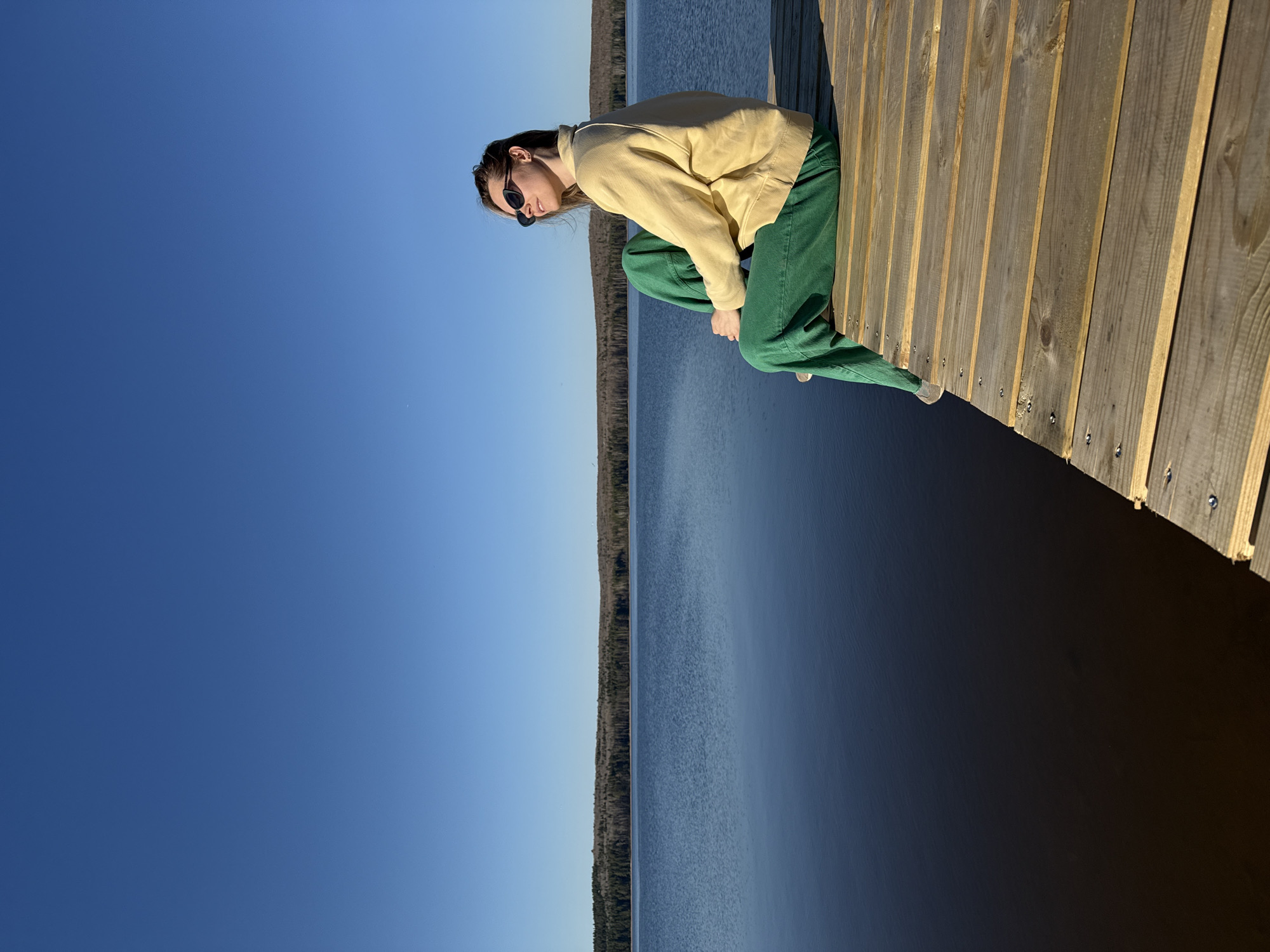 A young woman wearing sunglasses, a yellow sweater, and green pants sits on a wooden dock, looking down. The background shows a calm body of water and a treeline under a clear blue sky. The location is likely Chaikovsky, Russia.