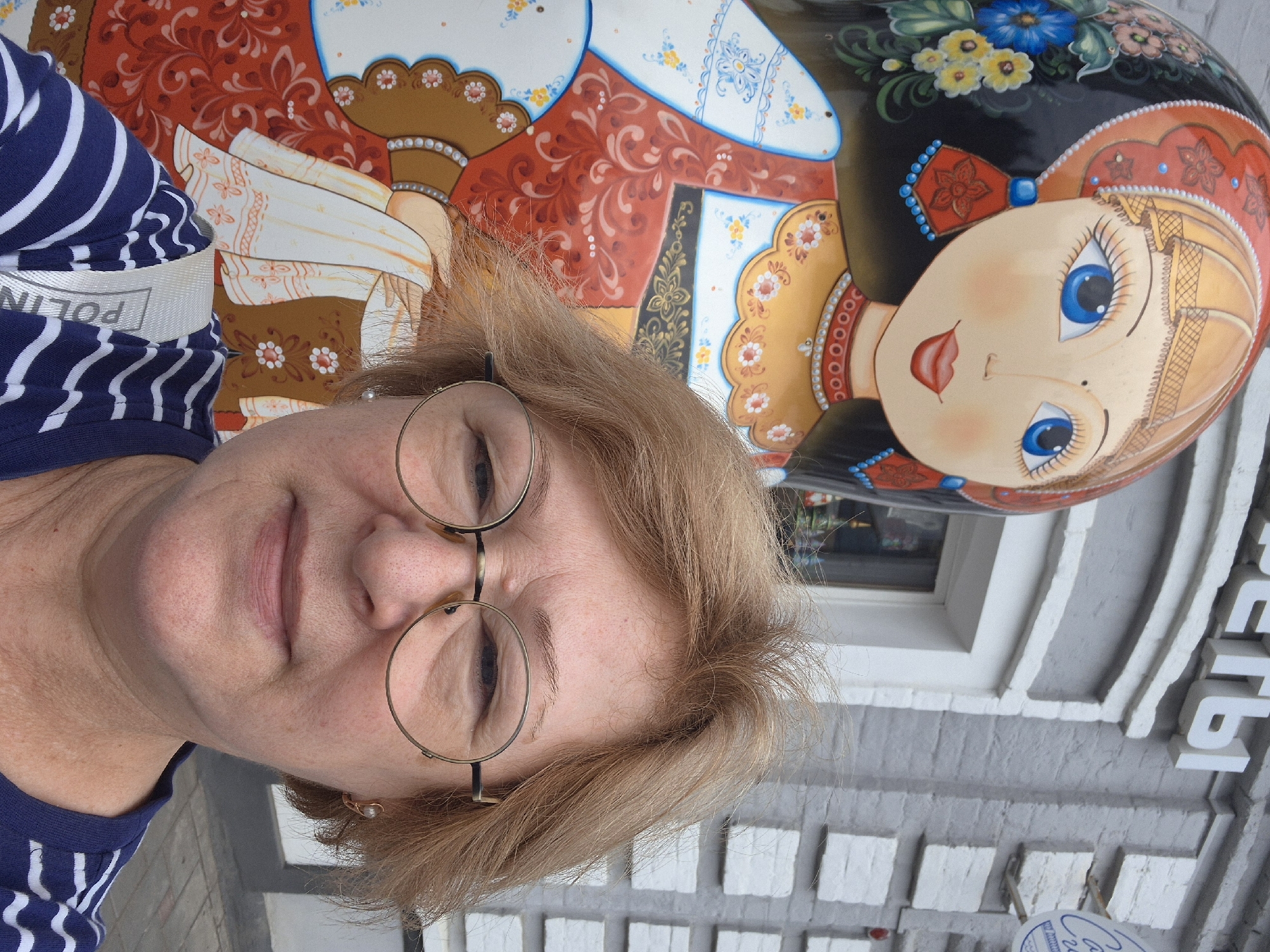 A medium shot selfie shows a woman with blonde hair and glasses standing in front of a large Matryoshka doll. The woman is wearing a blue and white striped shirt. The Matryoshka doll is elaborately painted with a woman's face and traditional Russian clothing. Behind them, a brick building with white trim is visible. The overall scene suggests an outdoor setting, possibly in a public area like a town square or street in Novokuybyshevsk, Russia. The perspective is from the woman's eye level, looking slightly upwards towards the doll.