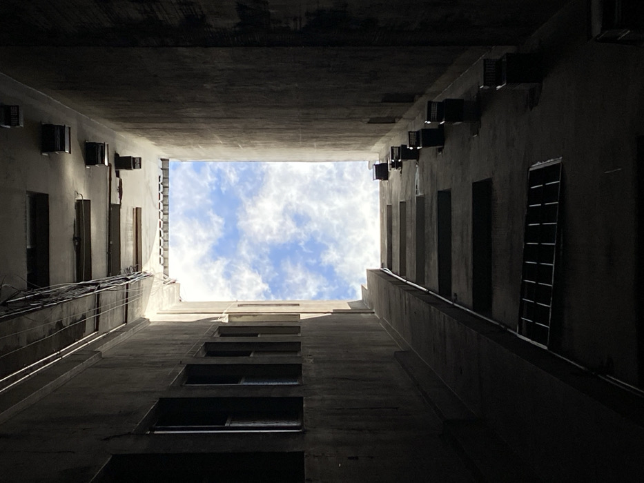 The image depicts an upward-looking view from the bottom of a narrow, multi-story urban lightwell or interior courtyard, characteristic of dense architecture found in Buenos Aires, Argentina. The scene is framed by three high walls of a grey, textured material, likely concrete, with their lower and middle sections heavily shadowed.
The left wall features multiple dark, rectangular window openings, vertically stacked, with a horizontal metal grate covering one lower opening. This wall also presents ascending, horizontal, step-like architectural ledges. Overhead, near the top of the left and back walls, utility cables are visible.
The right wall, also largely in deep shadow, displays a series of dark, rectangular ventilation units mounted vertically along its surface. At the very top, where the walls meet the sky, a horizontal band with small, repetitive architectural details is visible, suggesting a rooftop edge.
The only source of natural light is a rectangular opening directly above, revealing a bright blue sky filled with scattered white cumulus clouds. No individuals, animals, or dynamic actions are depicted within the static environment.