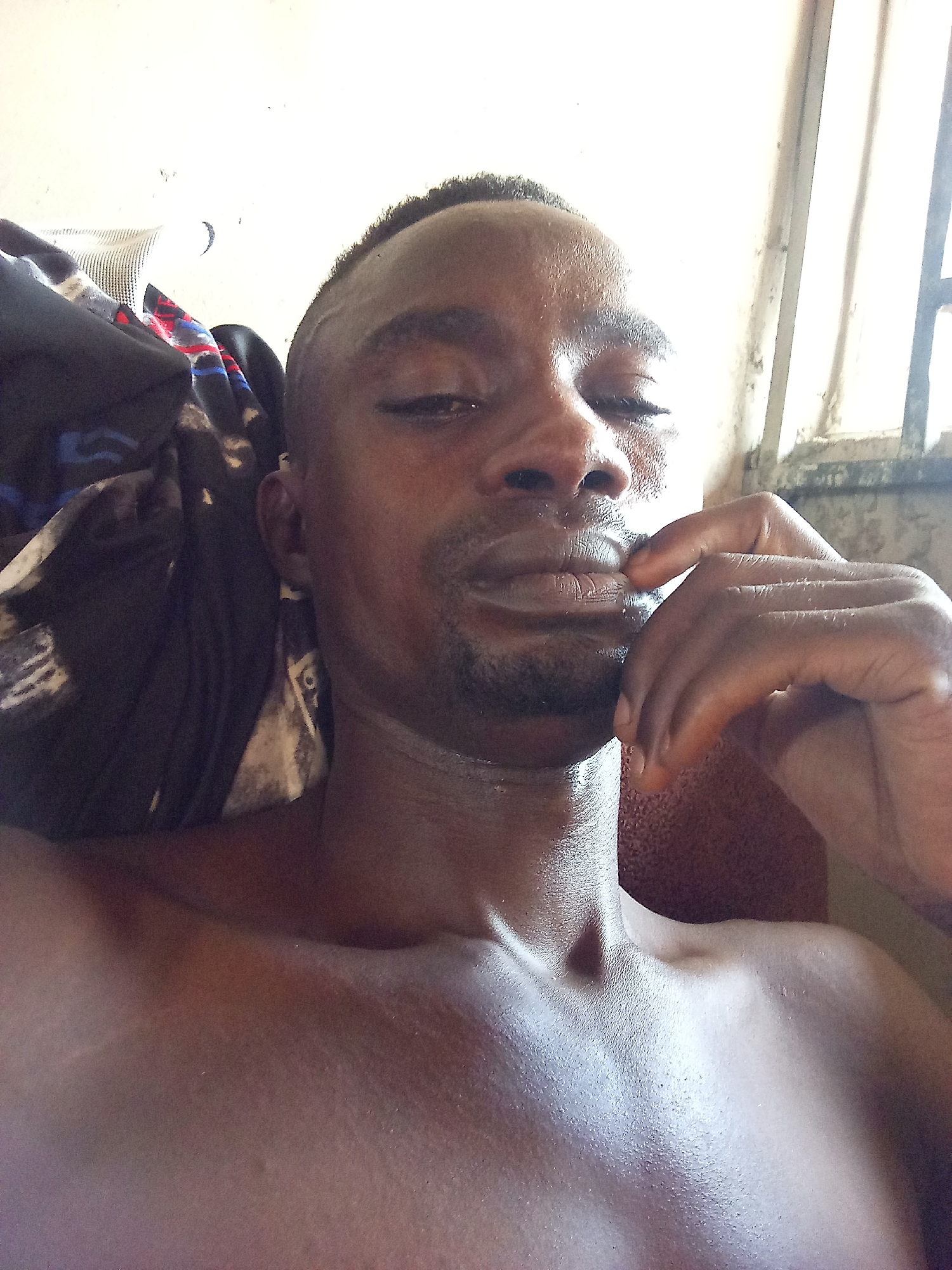 A close-up, eye-level selfie captures a young adult male with dark skin and a short, dark haircut. He is shirtless, revealing his bare chest and shoulders. His right hand is raised, with fingers gently touching his chin and lower lip, suggesting a pensive or contemplative mood. His eyes are partially closed or looking downward, and his lips are slightly parted. A subtle amount of facial hair is visible around his chin and upper lip.
The strong, bright natural light, likely from a window, illuminates his face and upper body, creating highlights on his forehead, nose, lips, and shoulders, making his skin appear glossy or dewy.
In the background, to the right, a window with vertical bars or a frame is visible, allowing bright light to enter. To the left, a pile of dark-colored fabric or clothing with some patterned elements is present. The walls appear light-colored. The overall setting appears to be indoors. No text is visible in the image. The scene is set in Adonkolo, Nigeria, during what appears to be daytime due to the bright natural light.