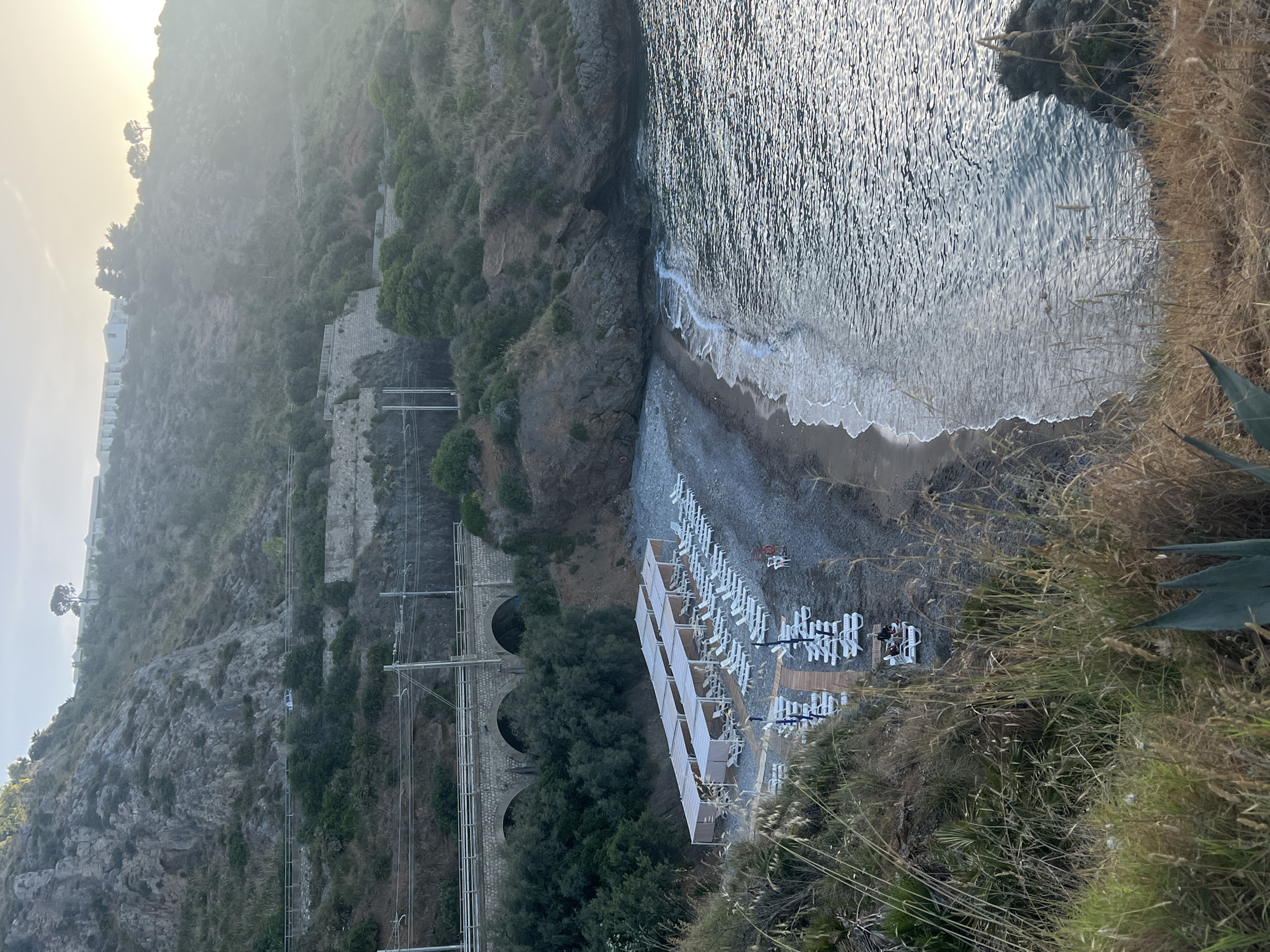 This image depicts a coastal cove in Sperone, Italy, viewed from an elevated perspective overlooking a pebble beach. The scene is illuminated by soft, hazy sunlight, suggesting either early morning or late afternoon.
In the upper portion, a steep, rocky mountainside dominates, covered with sparse green and brown vegetation. A winding road or path is visible traversing the slope, with a small, light-colored vehicle present on it. Higher up, a white building complex, likely a hotel or resort, is nestled among trees on the distant hilltop.
Mid-ground features a railway line running horizontally across the image, supported by overhead electrical infrastructure. Below the railway, a multi-arched stone viaduct spans a section of the terrain, partially obscured by dense green foliage.
The focus shifts to the beach in the lower-mid section, consisting of dark grey pebbles. Twelve rectangular sunshade structures, or pergolas with light-colored fabric tops, are arranged in two parallel rows. Numerous white plastic sun loungers are positioned beneath and around these pergolas; most appear unoccupied. A wooden boardwalk or pathway extends from the foreground onto the beach, leading towards the sunshades. One individual is discernible sitting on a sun lounger near the end of the boardwalk, partially hidden by foreground foliage. A red lifebuoy is mounted on a stand to the right of the loungers.
To the right, the calm, dark water of the sea laps the pebble shore with small white waves. The water surface reflects the overhead light, creating a shimmering effect. A rocky outcrop defines the right edge of the cove where it meets the sea.
The immediate foreground consists of dry, tall grasses and an agave-like plant, framing the panoramic view of the beach and sea below.