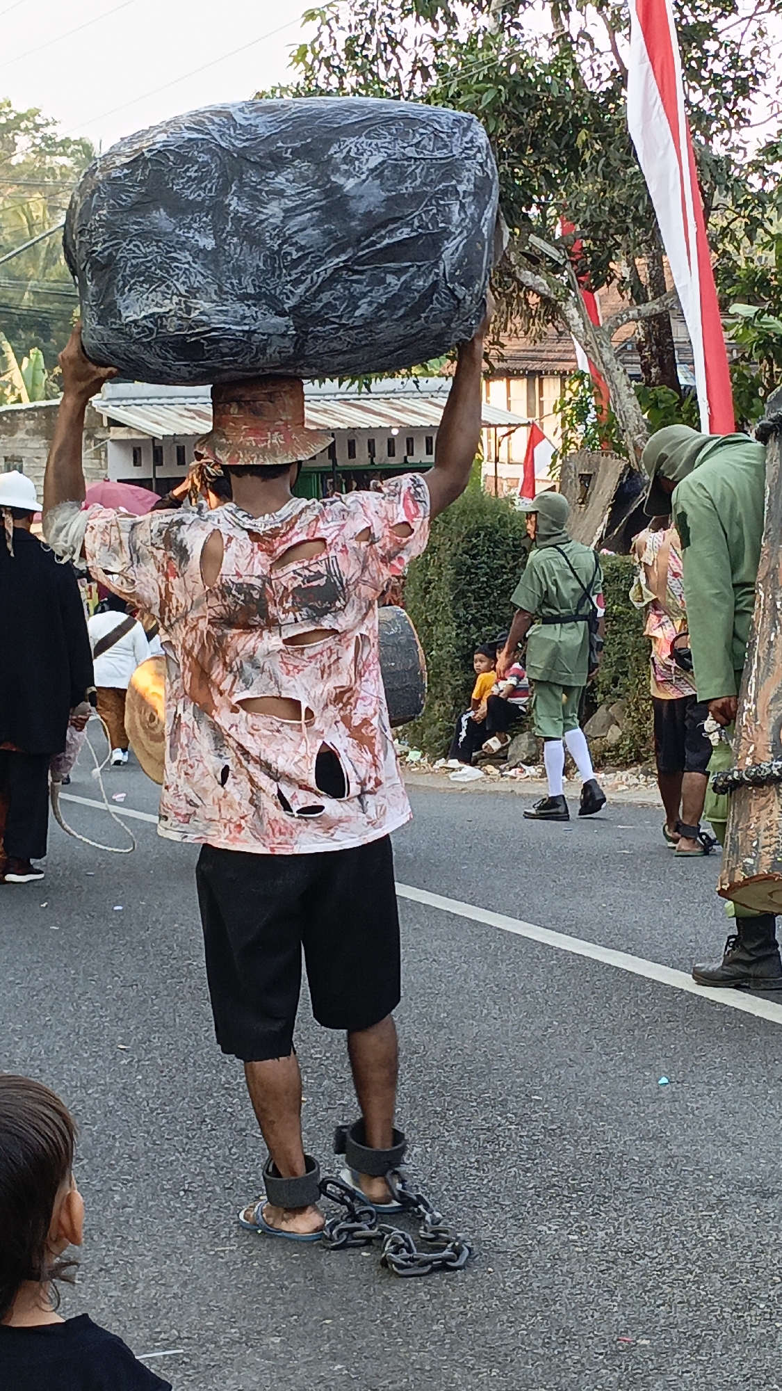 A man in Krajan Lor, Indonesia, is depicted from behind, carrying a large, grey, rock-like object on his head. He wears a torn, patterned t-shirt and black shorts, with heavy chains attached to his ankles. The scene appears to be a street procession or festival, indicated by the presence of other individuals in costumes and spectators. A red and white flag, possibly the Indonesian flag, is visible in the background, along with trees and buildings.