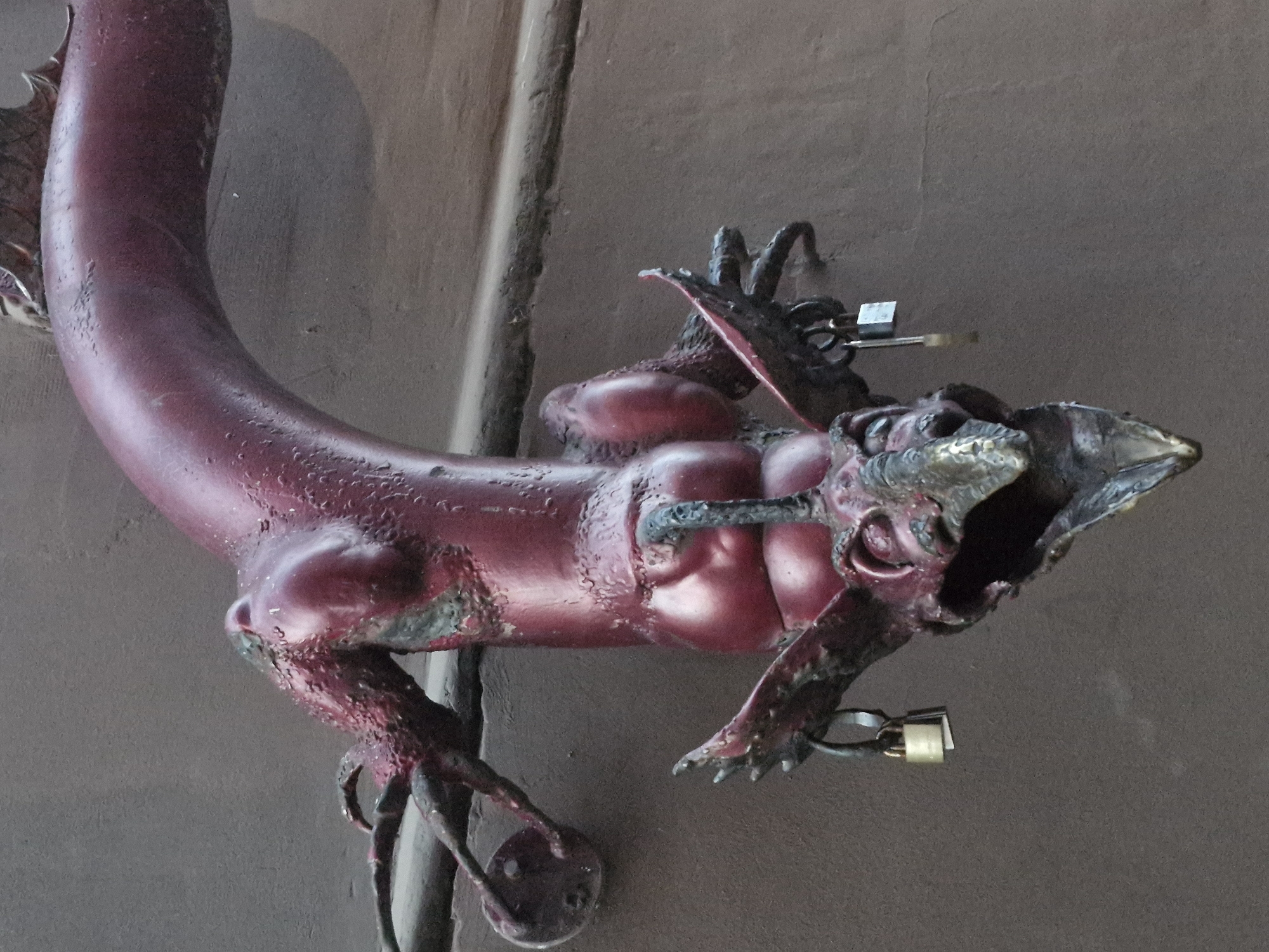 The image shows a close-up of a metallic sculpture of a mythical creature, possibly a dragon or gargoyle, painted in a reddish-purple hue. The sculpture is attached to a textured, dark brown wall. The creature is depicted hanging upside down, with its head facing downwards and its body curving upwards and to the right. Its head features prominent wings, sharp claws, and an open mouth revealing a pointed tongue and teeth. Two small brass padlocks are attached to the creature's body by metal loops. The left padlock hangs from a loop near its rear, and the right padlock hangs from a loop further up its body. Small droplets, possibly rain or moisture, are visible on the surface of the sculpture. The background wall appears to be made of rough plaster or concrete.
This sculpture is located in Klaipėda, Lithuania. Specifically, it is the "Sculpture of a Dragon" or "Dragon Gate" found in the Old Town of Klaipėda, near Theatre Square. The locks often represent pledges of love or commitment by couples.