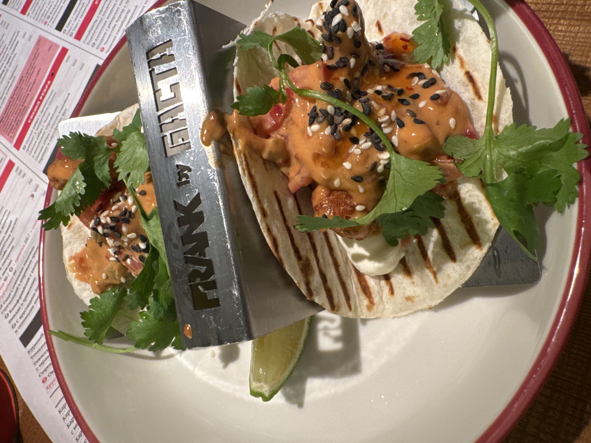 Here is a description of the image:
The photograph shows a plate of food, seemingly two tacos, placed on a table with a menu partially visible in the background. The tacos are arranged on a white plate with a red rim and are presented with a slice of lime. A metal serving utensil with the words "FRANK by FACTA" cut out of it is angled across the tacos. The tacos contain a filling with an orange-colored sauce, some visible ingredients, and a sprinkle of black and white sesame seeds. There is also some greenery in the form of herbs, likely cilantro, as garnishing. The setting appears to be a restaurant. It is located in Estosadok, Russia.
