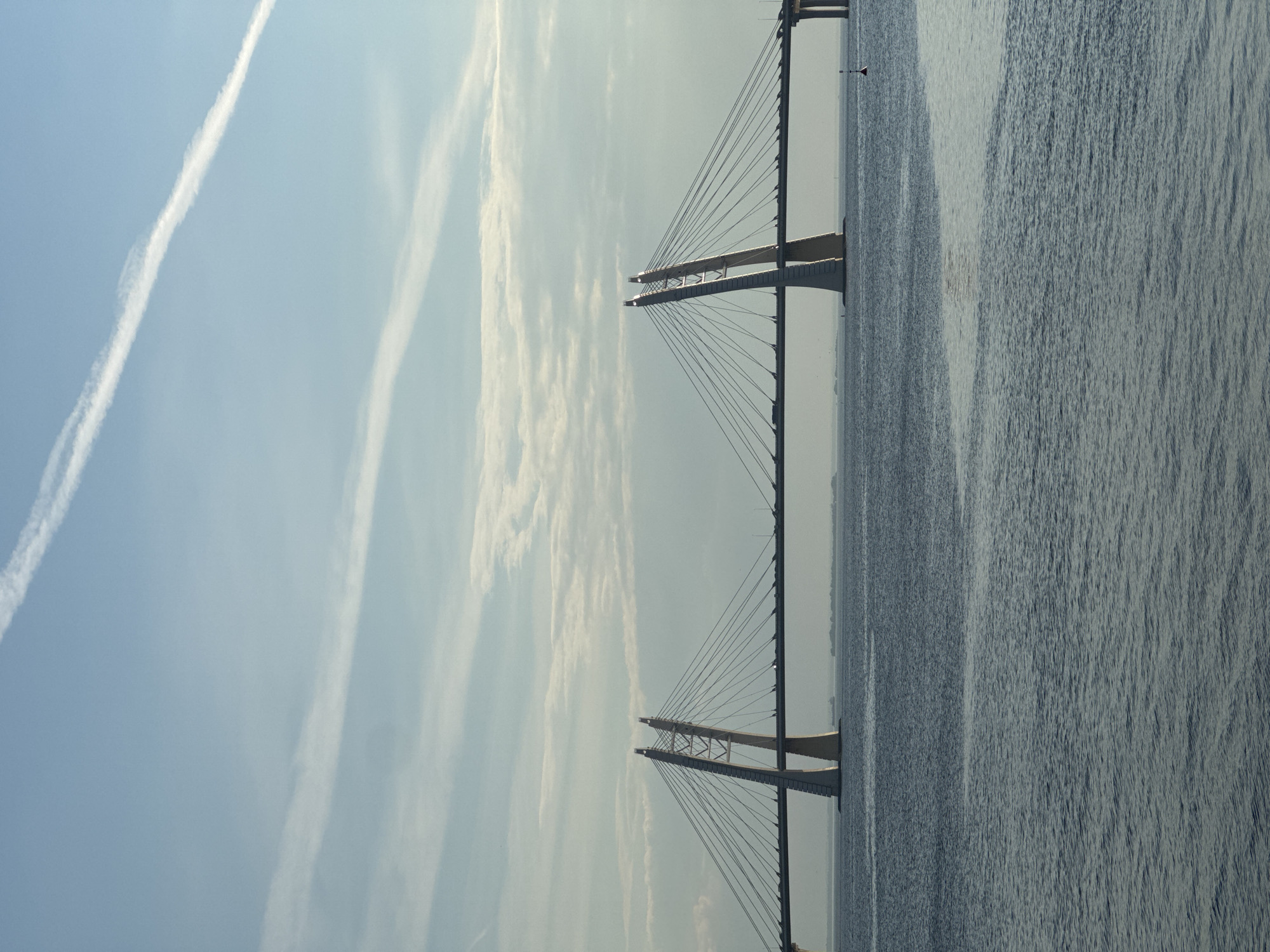 The image displays a vast outdoor scene featuring a modern cable-stayed bridge spanning a wide body of water, under a partially cloudy sky. The photograph, when correctly oriented, shows the bridge stretching horizontally across the middle ground. Two distinct, light-colored bridge towers, characterized by their A-frame structures, rise from the water, supporting numerous cables that fan out to hold the bridge deck.
The sky, occupying the upper portion of the frame, is a light blue, adorned with wispy white clouds and several long, thin contrails, suggesting airplane activity. The lighting indicates a bright day, possibly late morning or early afternoon, with clear visibility. Below the bridge, a large expanse of dark, rippled water fills the lower and right sections of the image, showing texture from subtle waves or reflections. A small, dark buoy with a reddish top is visible on the water further out from the bridge. In the distance, a faint shoreline or landmass can be seen on the horizon, beneath the bridge deck.
The scene highlights a marvel of modern engineering against a serene natural backdrop. This extensive structure, situated in Saint Petersburg, Russia, is likely a part of the city's Western High-Speed Diameter. No text is visible in the image.