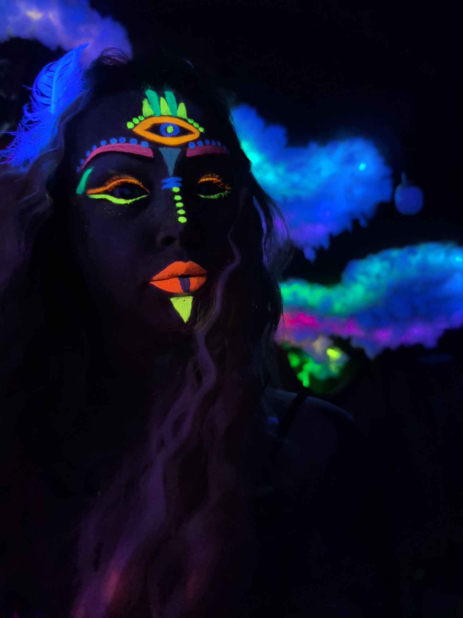 A person is depicted in a dark, blacklight-illuminated environment, showcasing elaborate glow-in-the-dark face paint and makeup. The subject, likely female with long, wavy hair, is positioned facing forward with a neutral expression. Her face is largely obscured by shadow, but vibrant, neon colors glow intensely under ultraviolet light.
Notable features include a prominent "third eye" design on the forehead, comprising an orange eye shape outlined in green with green flame-like elements above. Below this, an arc of pink dots and lines extends across the forehead, framing the eyes. The eyelids and surrounding areas are adorned with bright neon green and orange lines and dots, creating intricate patterns. The lips are painted a vivid orange with a vertical green stripe in the center and a striking neon green triangle directly beneath the lower lip. A series of small green dots extends down the nose.
To the left of the subject, blue, glowing feather-like elements are visible near her head, possibly part of a headpiece. The background features ethereal, cloud-like formations illuminated in shades of blue, green, pink, and purple, contributing to a surreal and artistic atmosphere. These elements suggest a possible art installation, a performance, or a themed event. The scene evokes a mysterious and vibrant mood, characteristic of a blacklight party, club, or creative photoshoot taking place at night or in a darkened indoor setting in Noviy, Russia. No visible text is present.